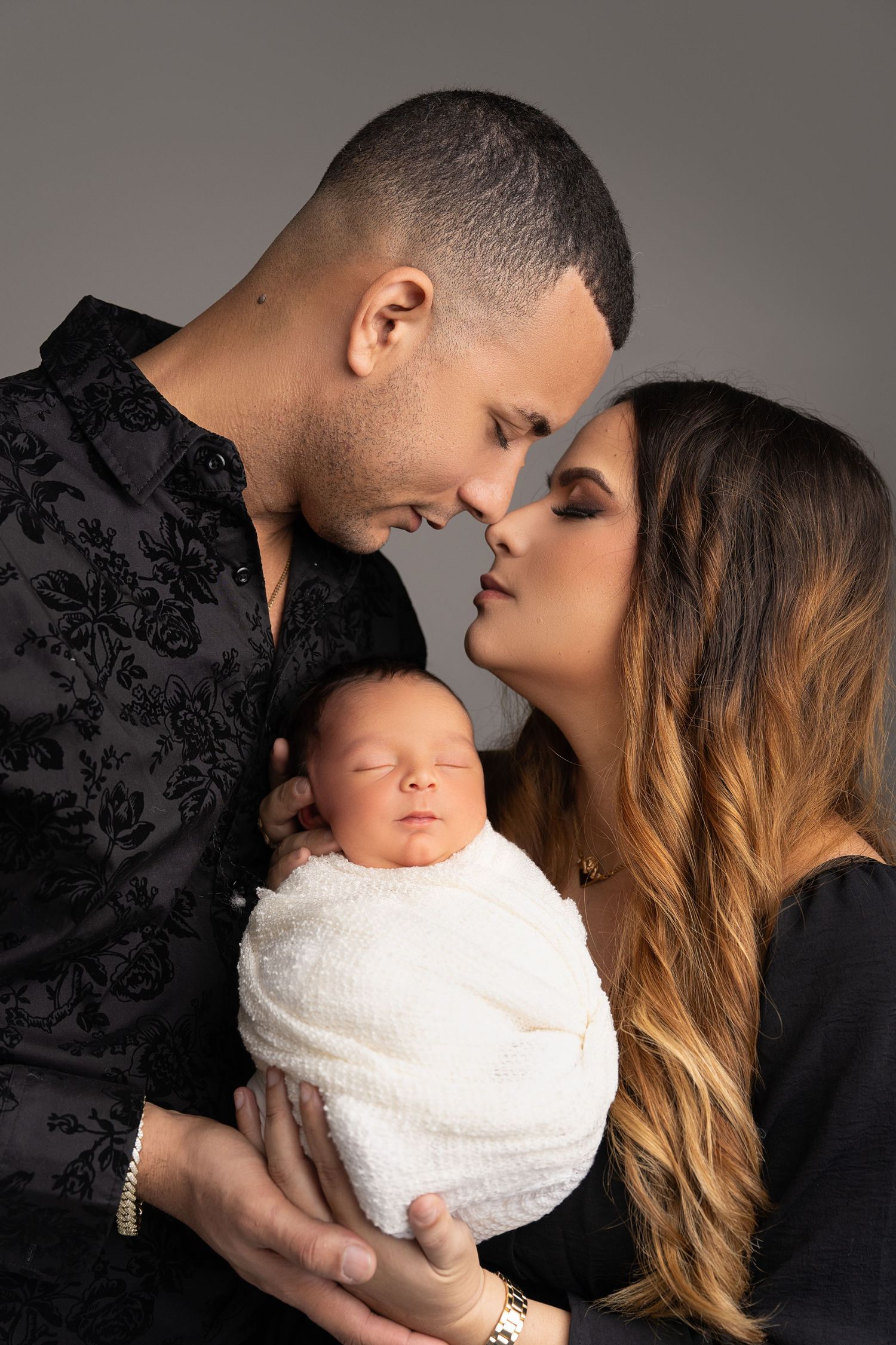 Intimate family portrait with sleeping newborn in white wrap against grey backdrop.