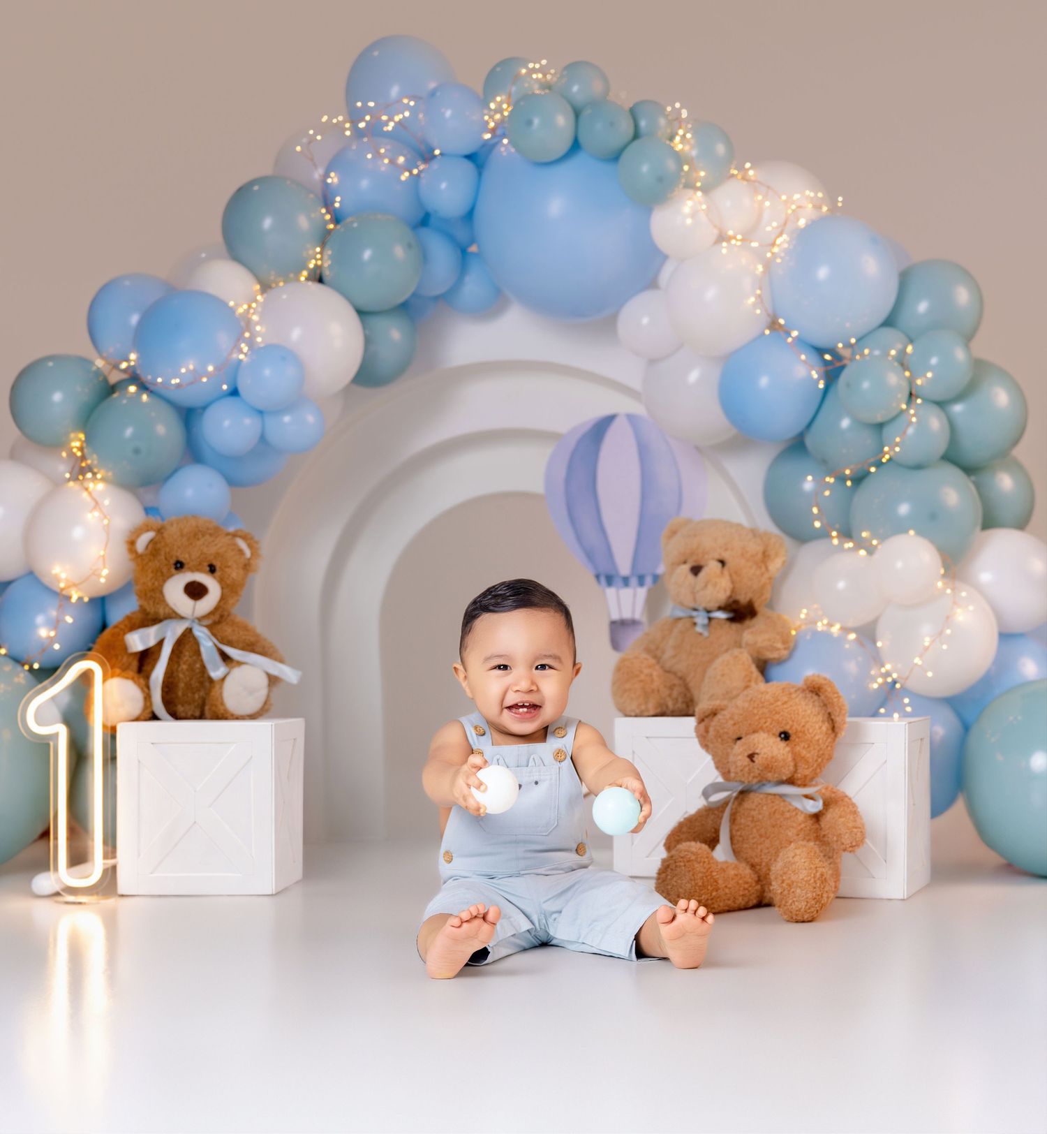 Baby boy sits smiling at his first birthday party with teddy bears, a number one prop, and a blue balloon arch.