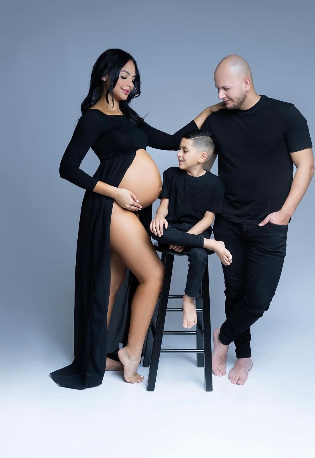 Pregnant woman in black maternity gown poses with husband and young son for a family maternity photo shoot.