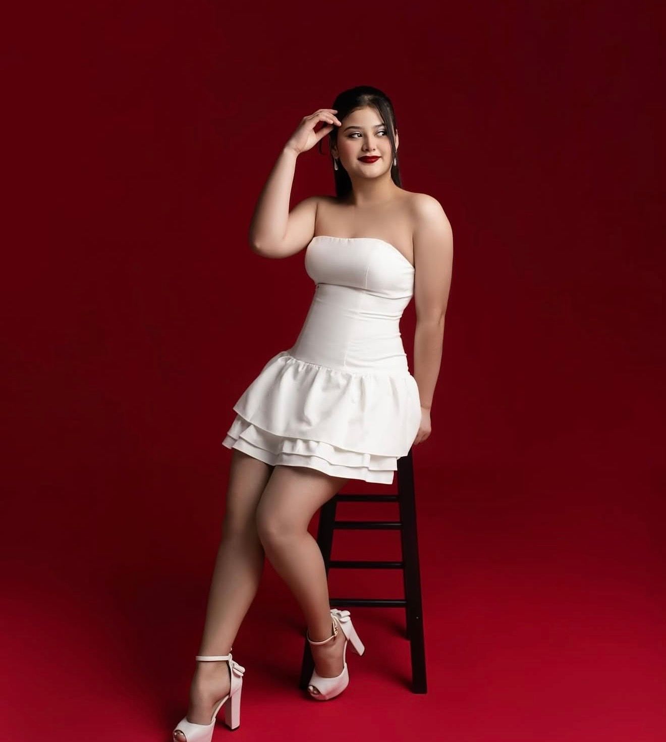 Woman in white strapless ruffled mini dress and heels poses on stool against bold red background.