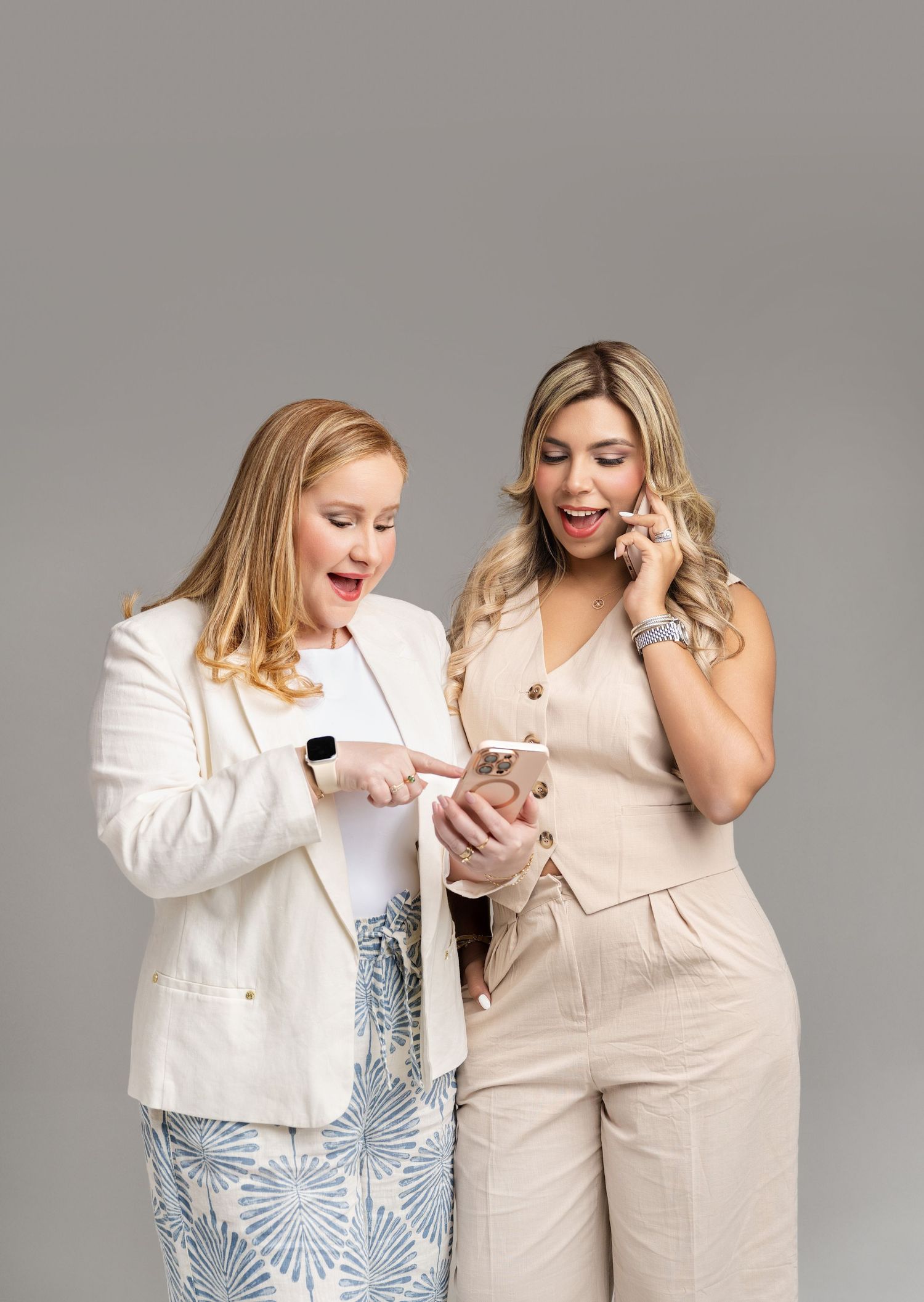 Two women reacting with surprise while looking at a smartphone, dressed in elegant business casual outfits on gray background.