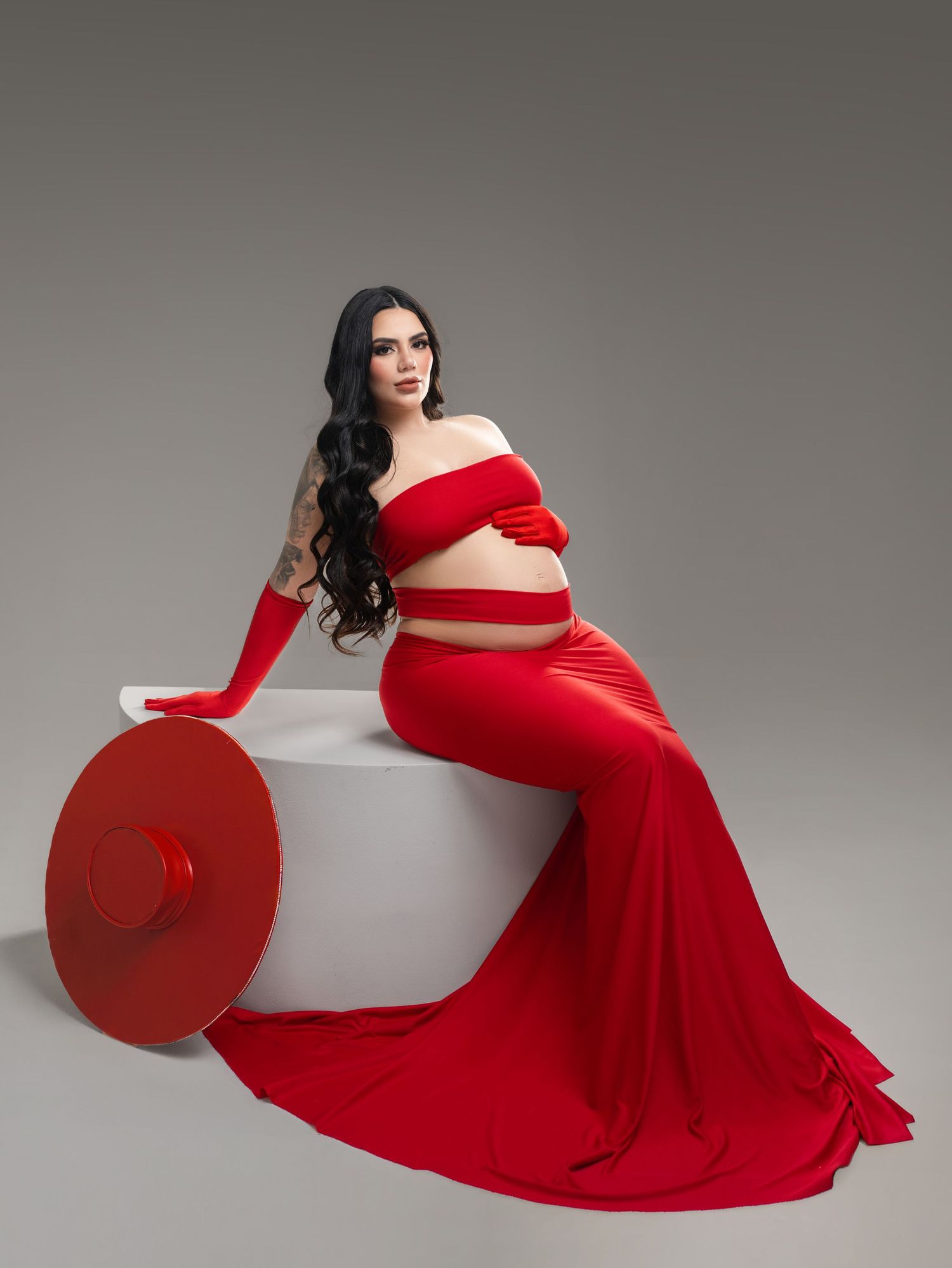 Pregnant woman in red off-shoulder crop top and flowing skirt sits gracefully on white cube with red hat prop.
