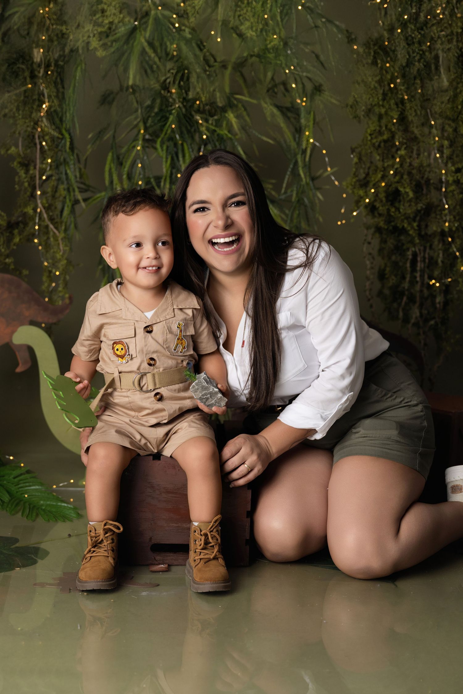 A smiling woman poses with a toddler boy in a safari outfit amid a jungle-themed backdrop with fairy lights and dinosaurs.