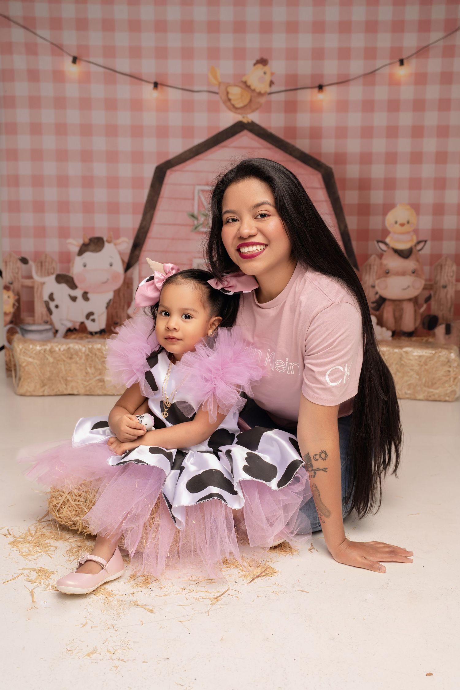 A woman and toddler girl pose in matching pink cow-print tutu dresses at a farm-themed photo shoot.