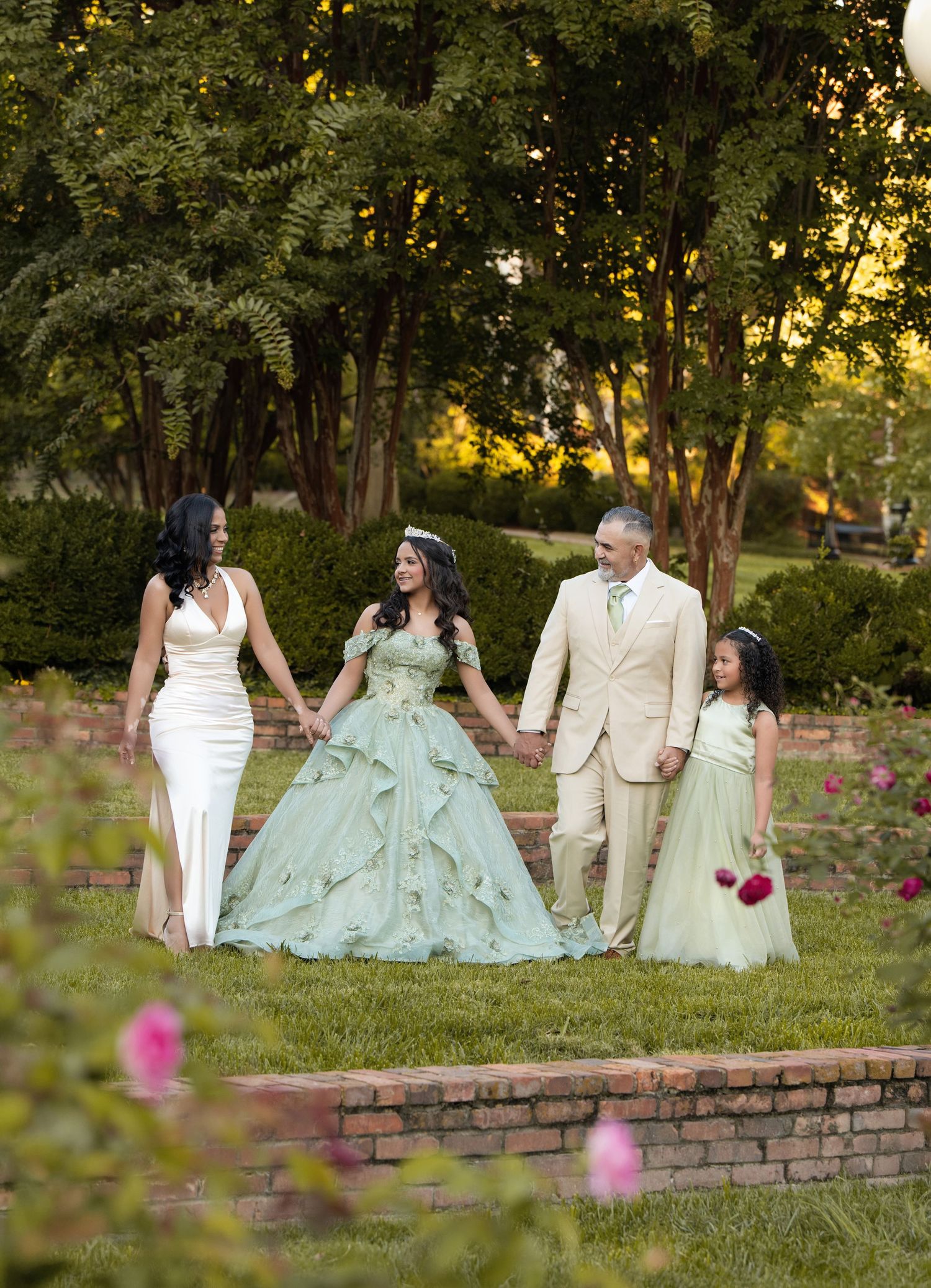 Family of four poses in garden during quinceañera celebration, girl in green ballgown holds hands with parents and sister.