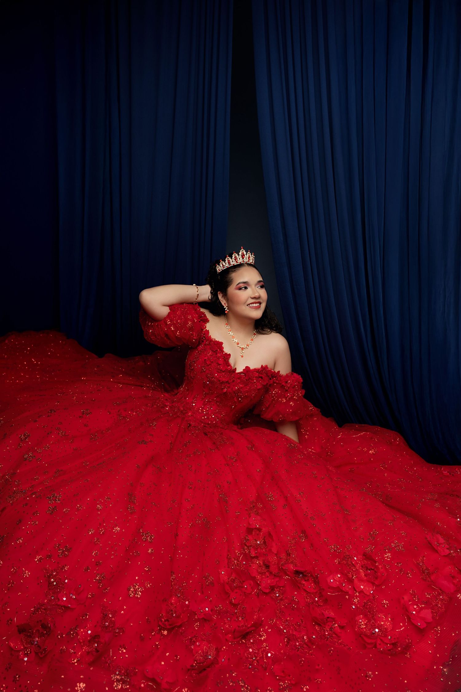 Woman in red ballgown with crown poses elegantly against blue curtains in a stunning quinceañera portrait.