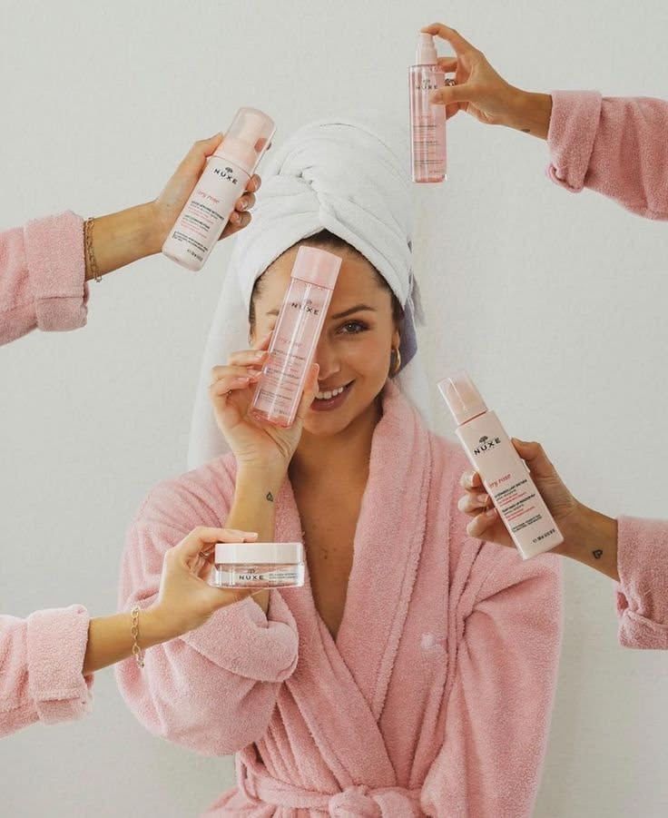 Woman in pink robe and towel turban surrounded by hands holding Nuxe skincare products.