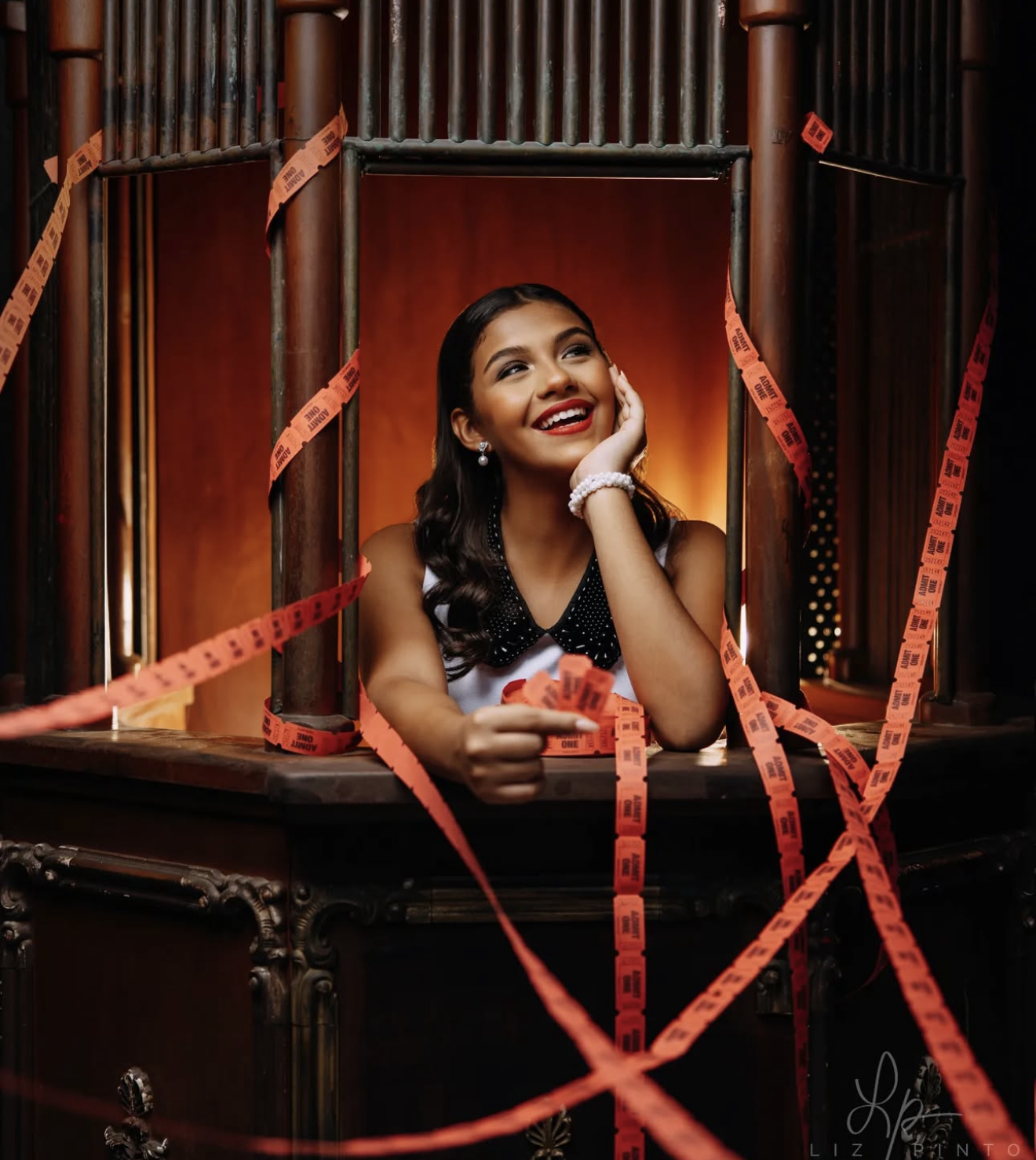 Young woman in elegant dress smiles joyfully inside a wooden cabinet surrounded by red ticket tape streamers.