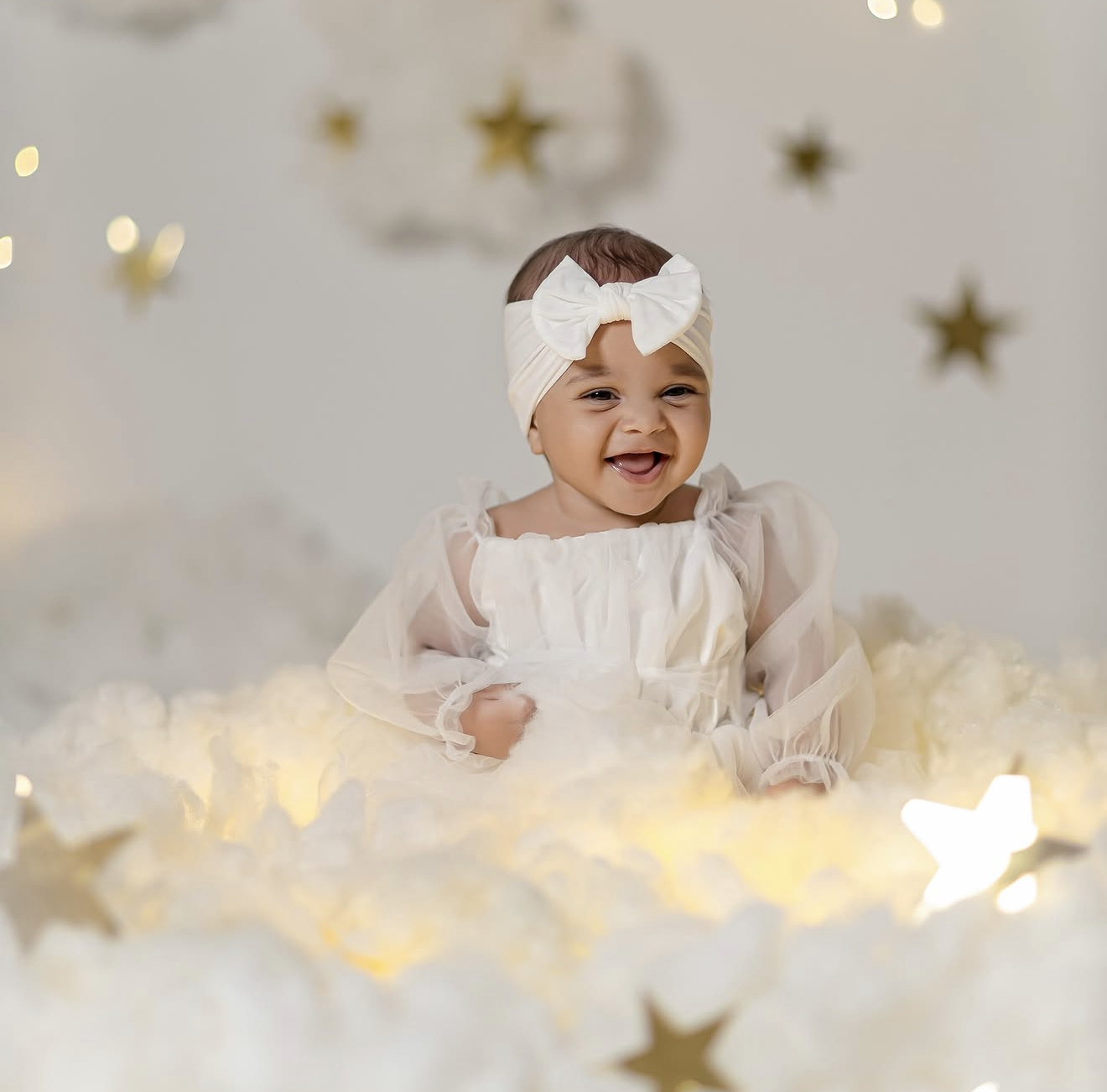 Smiling baby in white dress and bow headband sits among glowing lights and gold stars in a dreamy photo shoot.