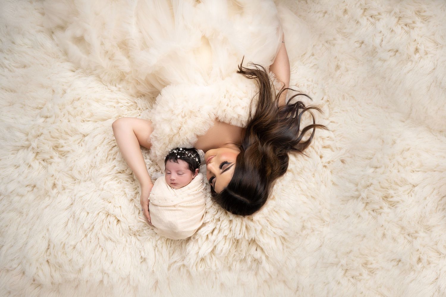 Aerial view of a mother in a white dress lying on a fluffy cream rug with her swaddled newborn baby.