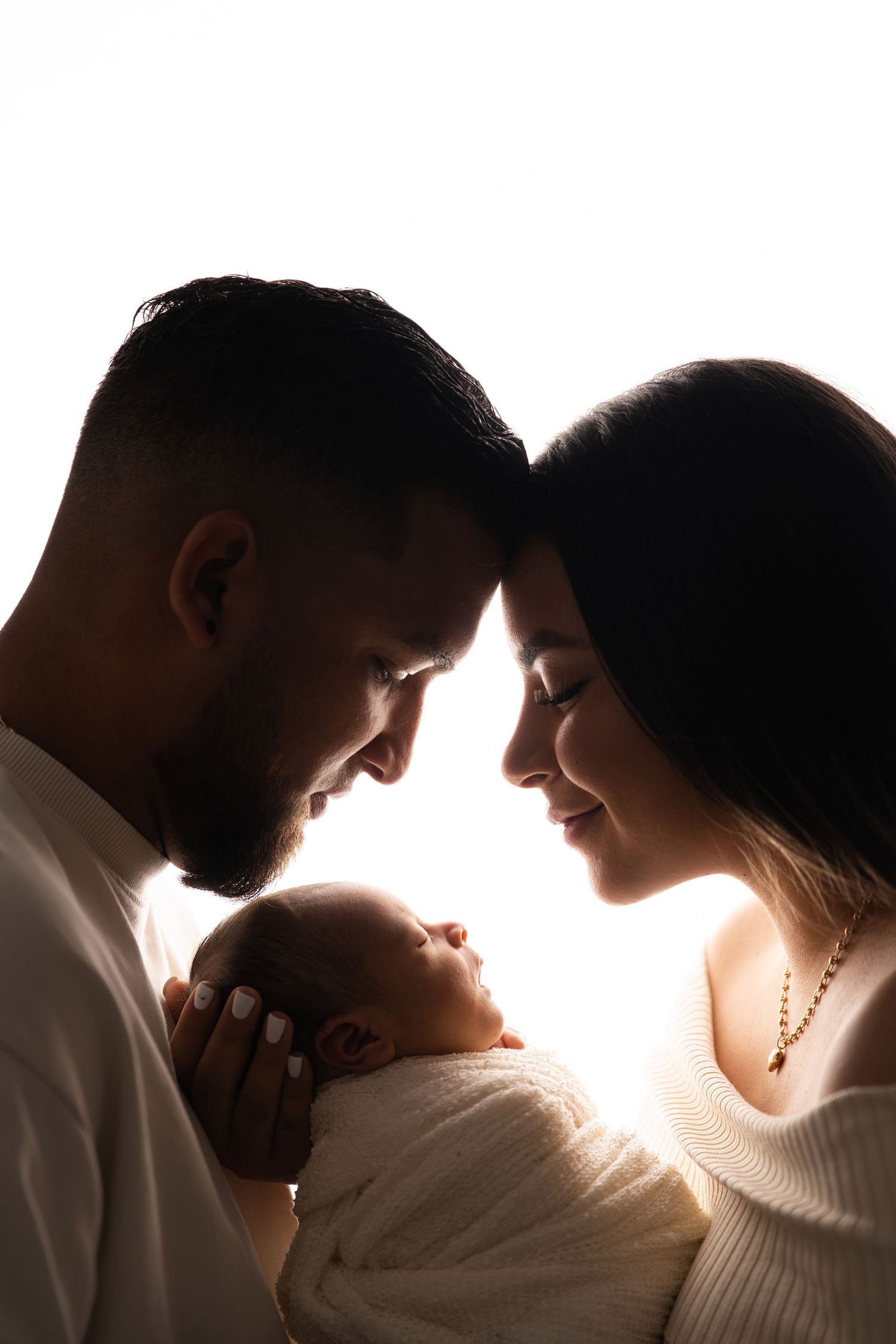 Silhouette of parents touching foreheads while lovingly holding their sleeping newborn baby against bright white background.
