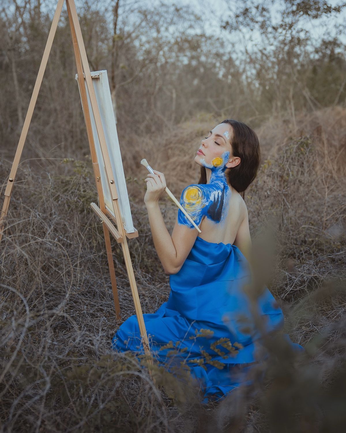 Woman in blue dress painting on canvas easel outdoors, with paint on her hands and face, surrounded by dry grass.