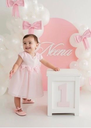 Baby girl in pink dress smiles at first birthday party with balloon arch and name sign reading Neena.