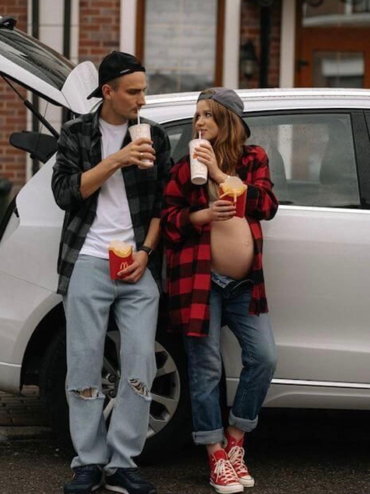 Pregnant couple leaning on car enjoying McDonald's milkshakes and fries outdoors.