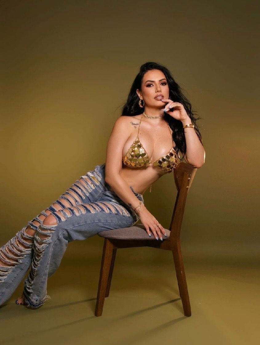 Woman in gold bikini top and ripped jeans posing on a wooden chair against a golden background.