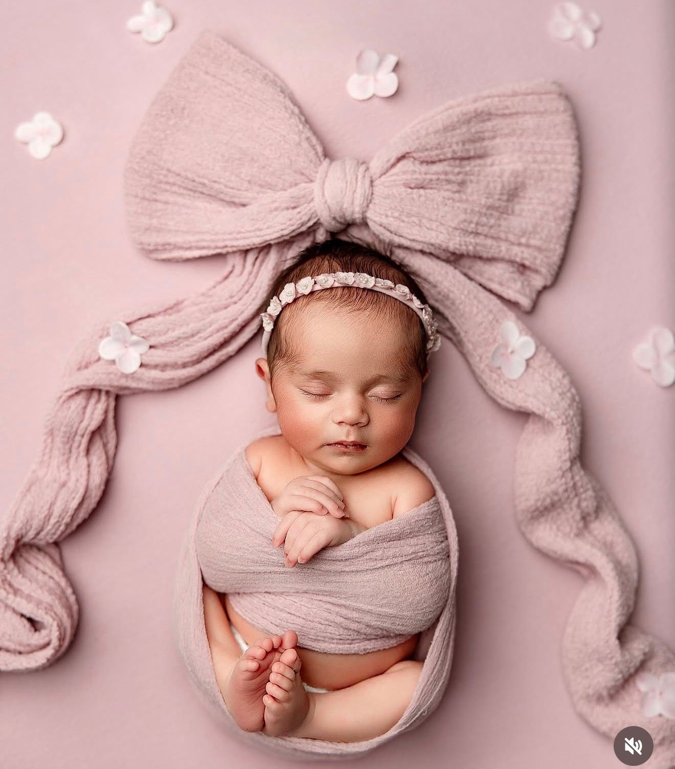 Newborn baby girl wrapped in pink knit fabric with a large bow, sleeping peacefully on a pink background.