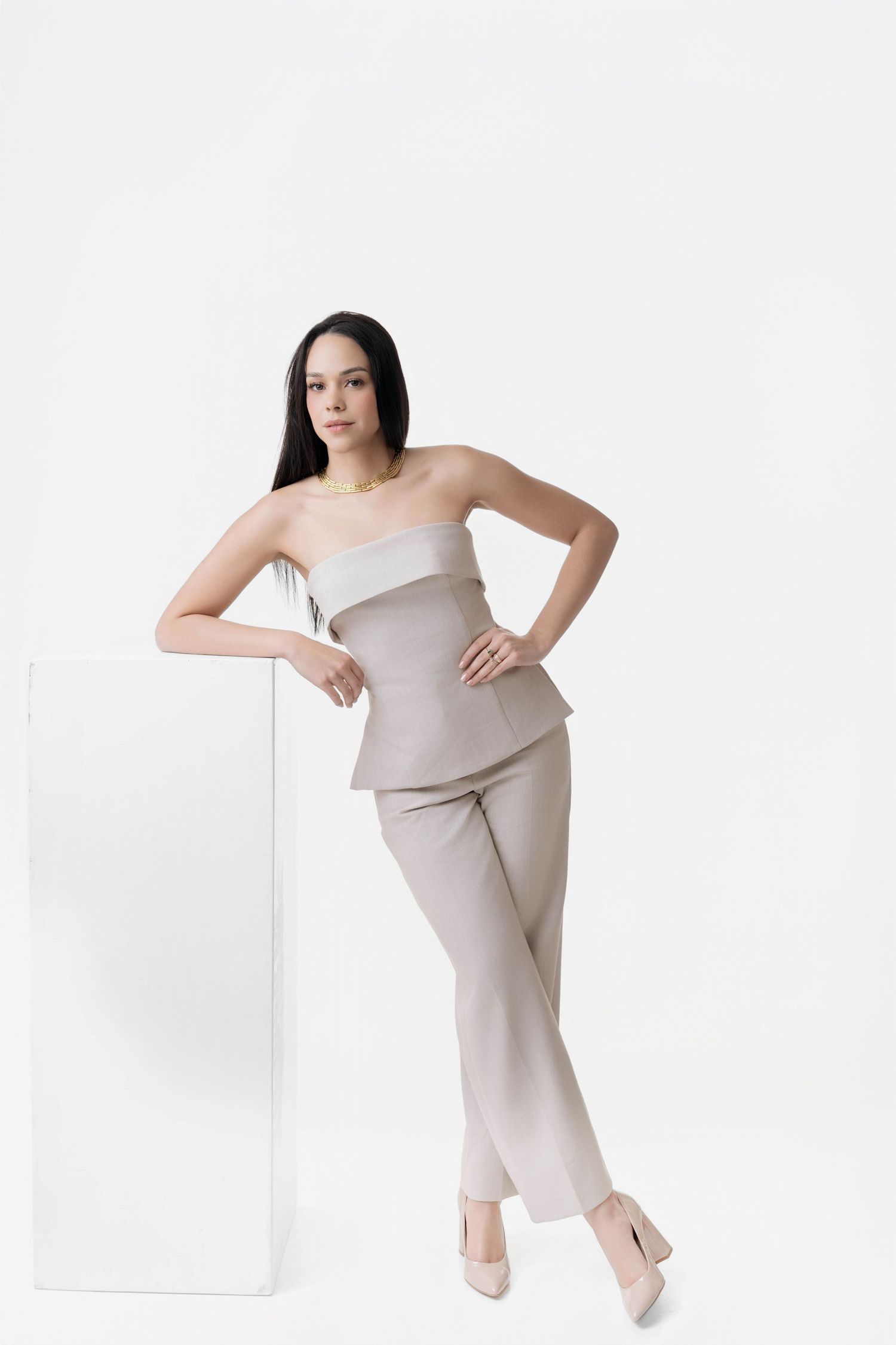 Woman with long dark hair poses leaning on a white pedestal in a taupe strapless peplum top, matching trousers and nude pointed heels.