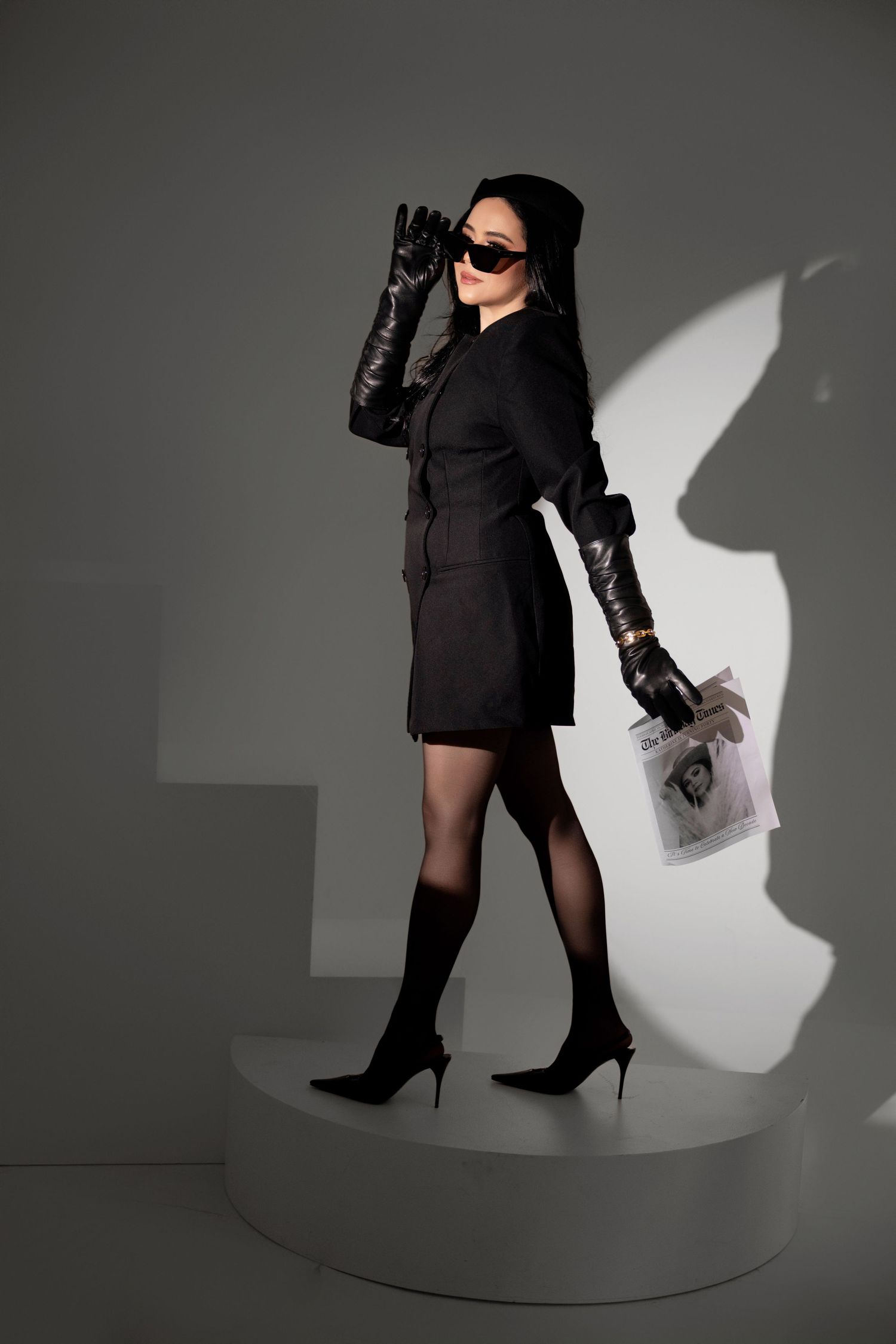 Woman in black dress, gloves, sunglasses, and heels poses on white platform against gray background.