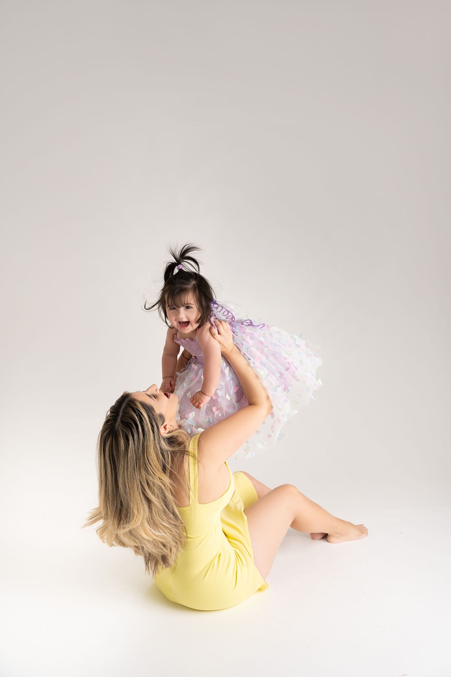 A woman in yellow dress lifts a laughing toddler in purple tutu dress overhead while sitting on white floor.