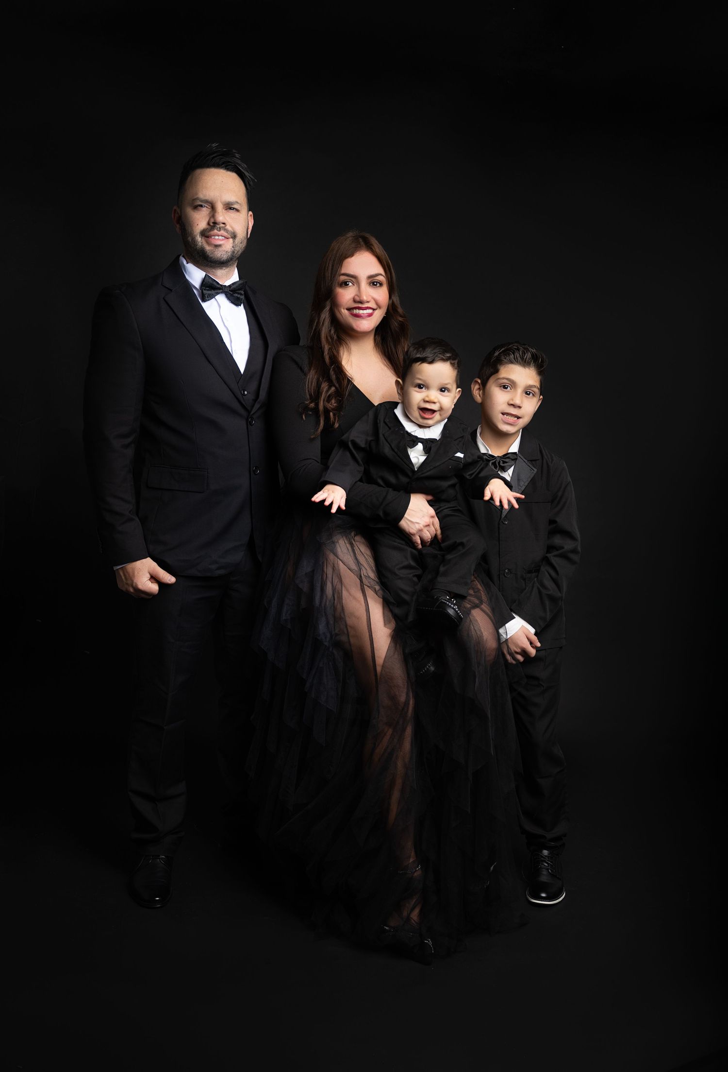 Family of four in elegant black formal wear posing together against a dark studio background.
