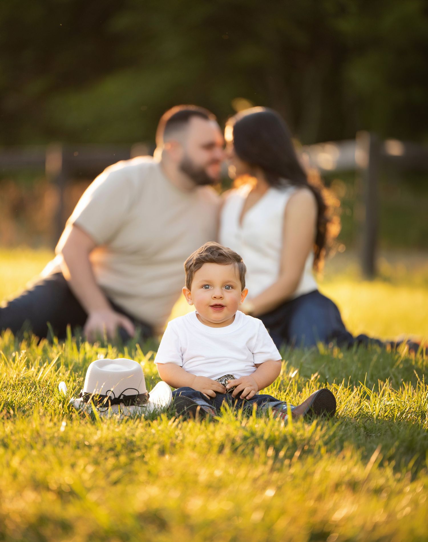 A baby boy sits on green grass smiling while his parents share a kiss behind him in a sunny park.