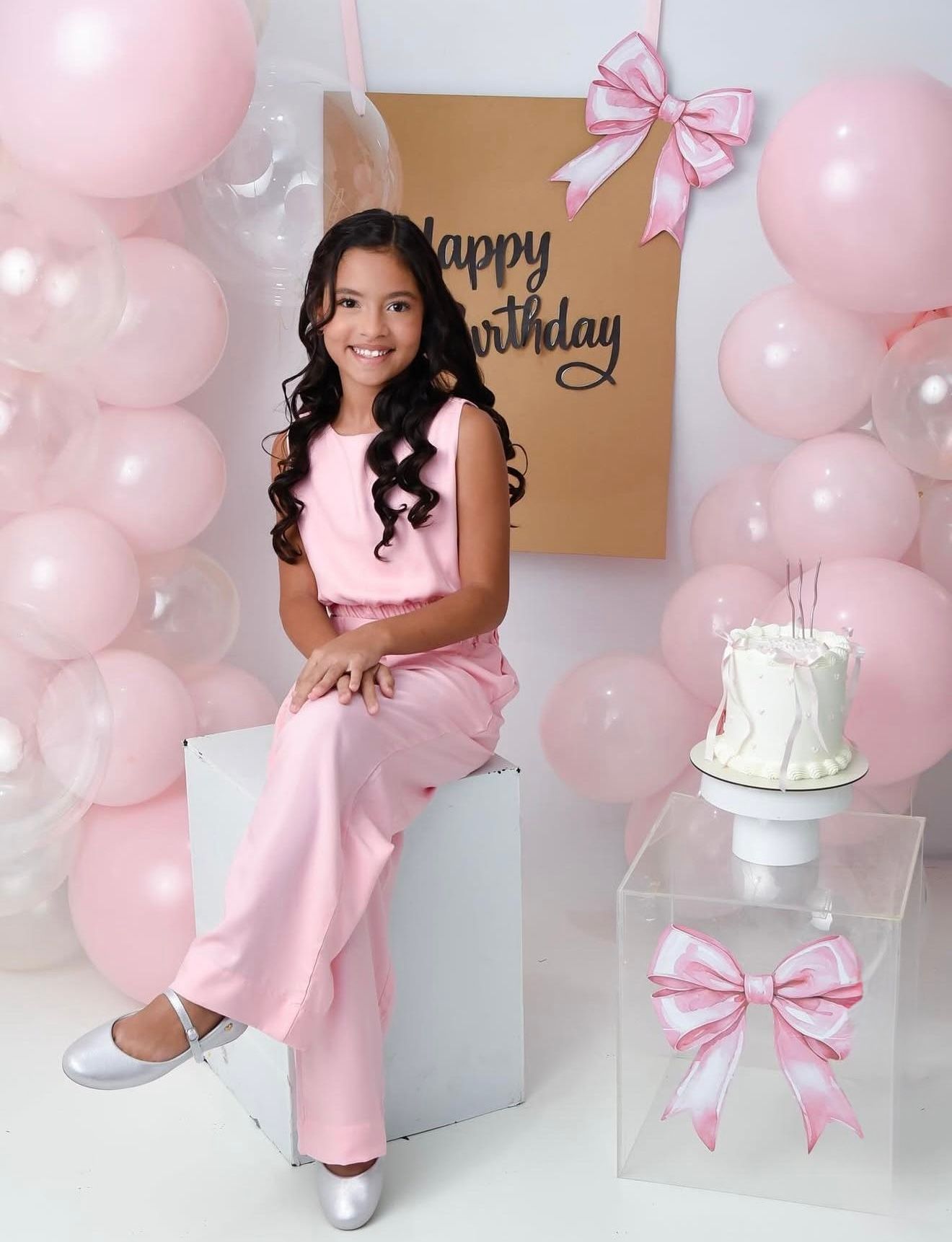 Young girl in pink outfit sitting by birthday cake with pink balloons and Happy Birthday sign backdrop.
