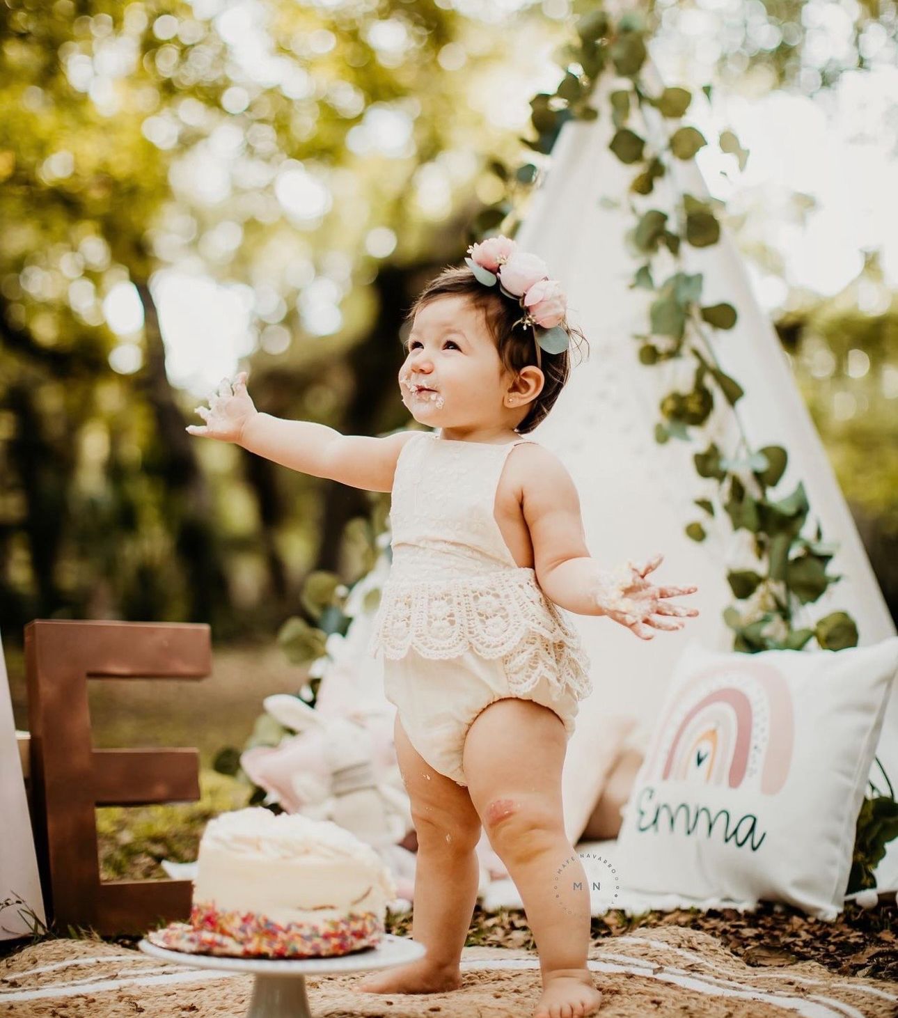 Baby girl in lace romper and floral headband stands by first birthday cake near a boho teepee outdoors.