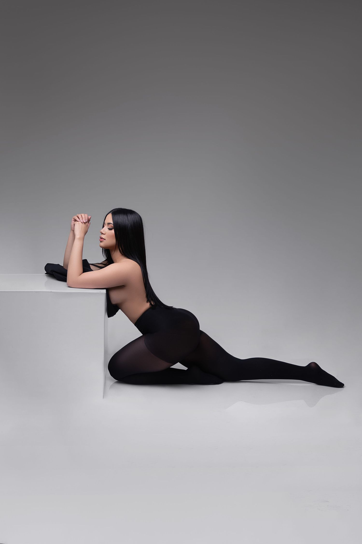 A woman in black bodysuit and tights poses gracefully beside a white cube on a gray studio background.