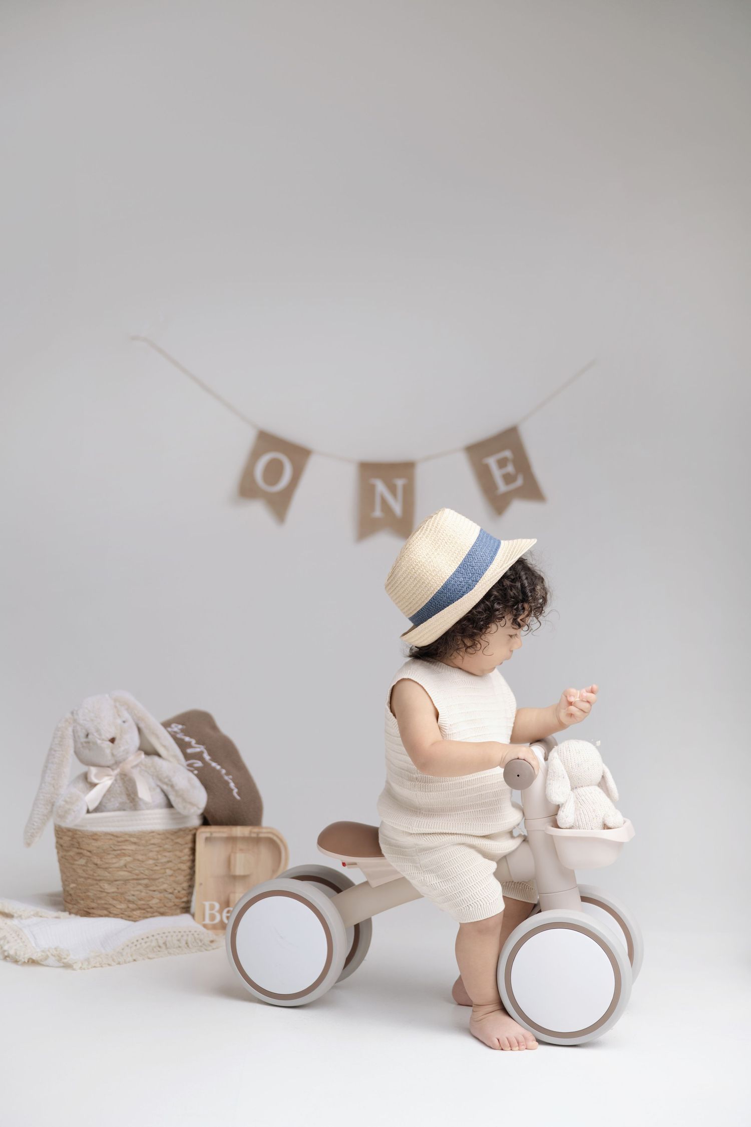 A toddler in a straw hat rides a wooden balance bike beside a wicker basket with bunny toys under a ONE birthday banner.