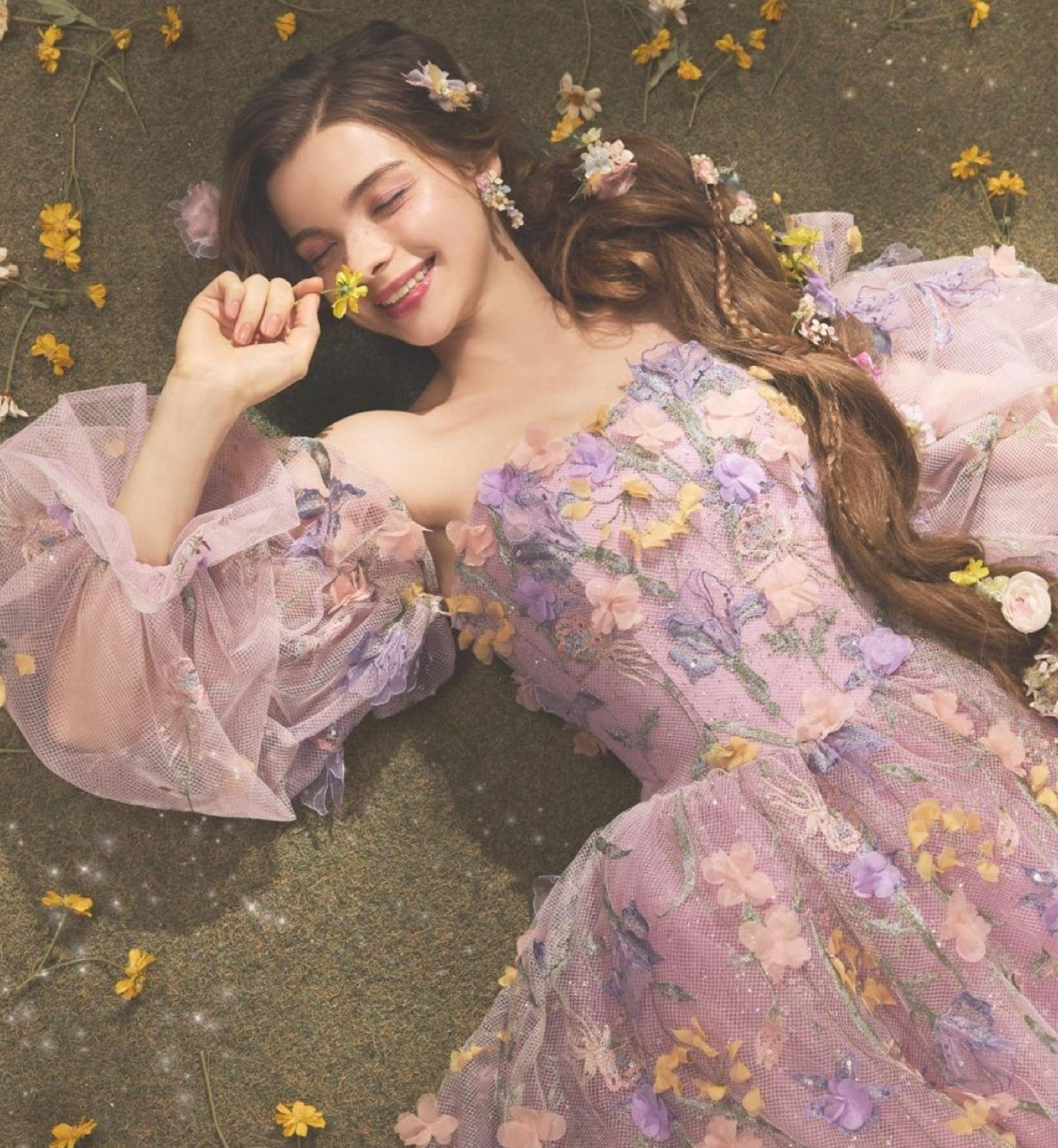 Young woman in floral lavender dress lying among flowers, smiling dreamily with flowers in her long brown hair.