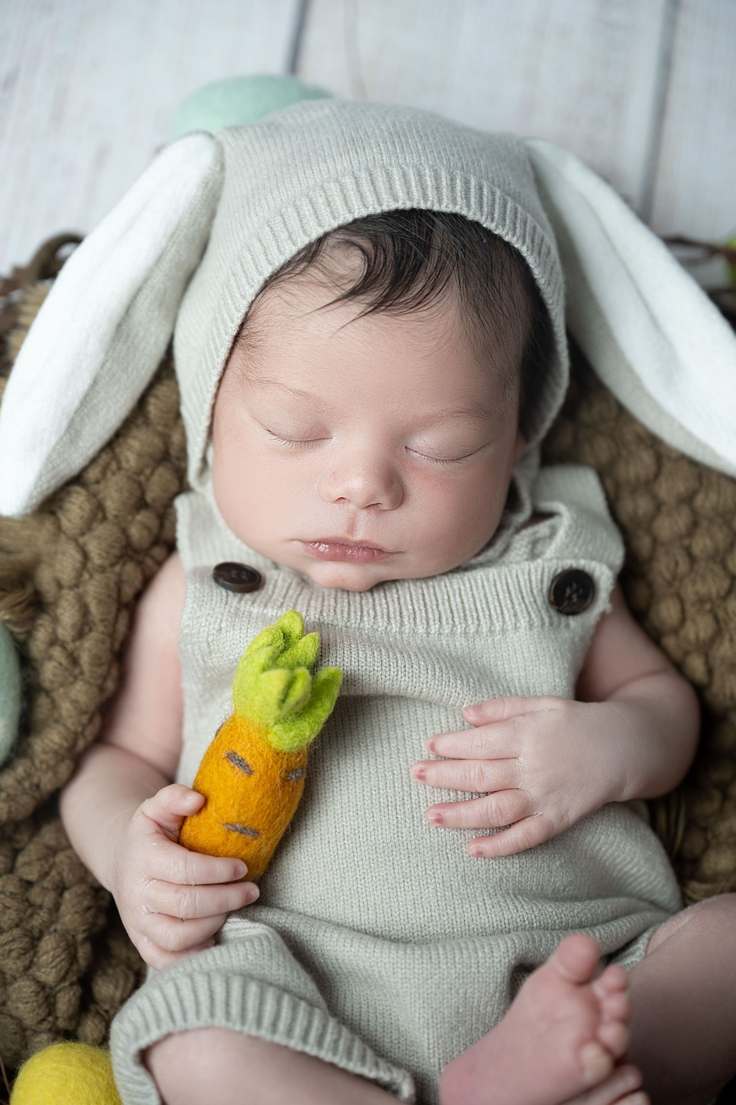 Sleeping newborn in gray bunny outfit holds plush carrot toy while resting on brown knit blanket.