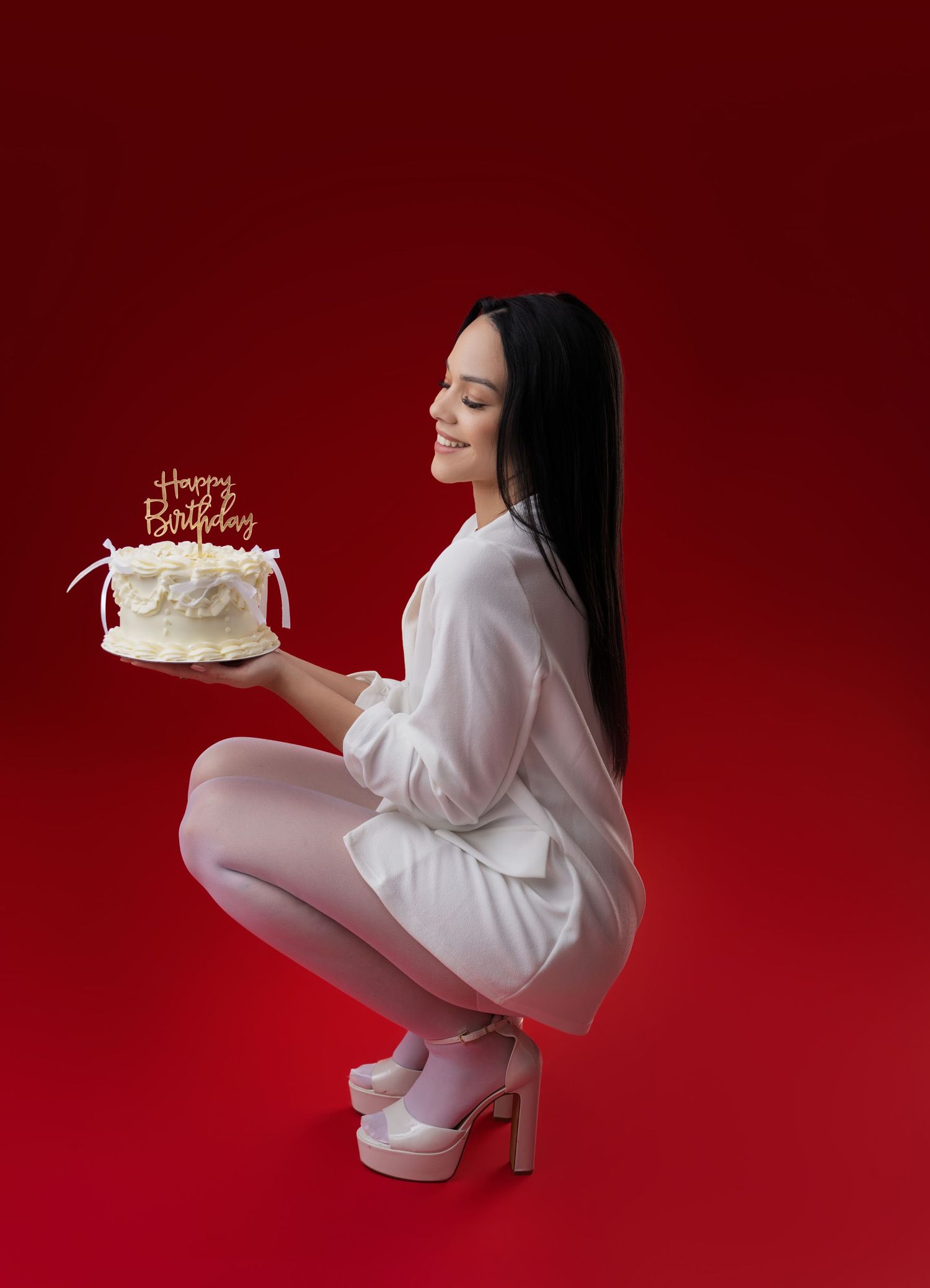 Smiling woman in a white outfit and platform heels crouches against a red backdrop, holding a white Happy Birthday cake.