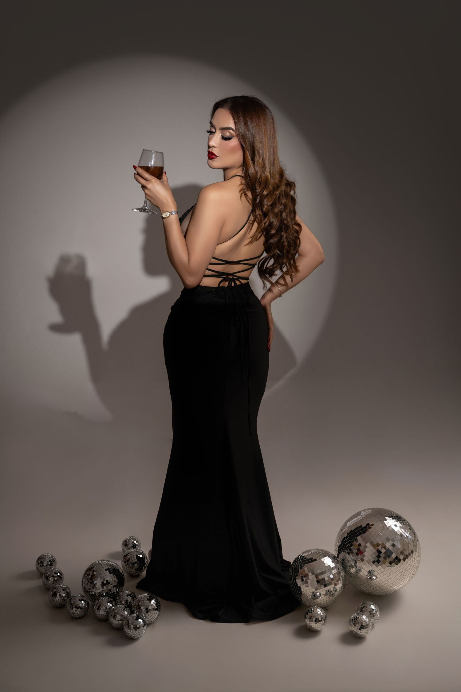 Woman in an elegant black backless evening gown holding a wine glass, posed beside silver disco balls with a dramatic spotlight shadow.