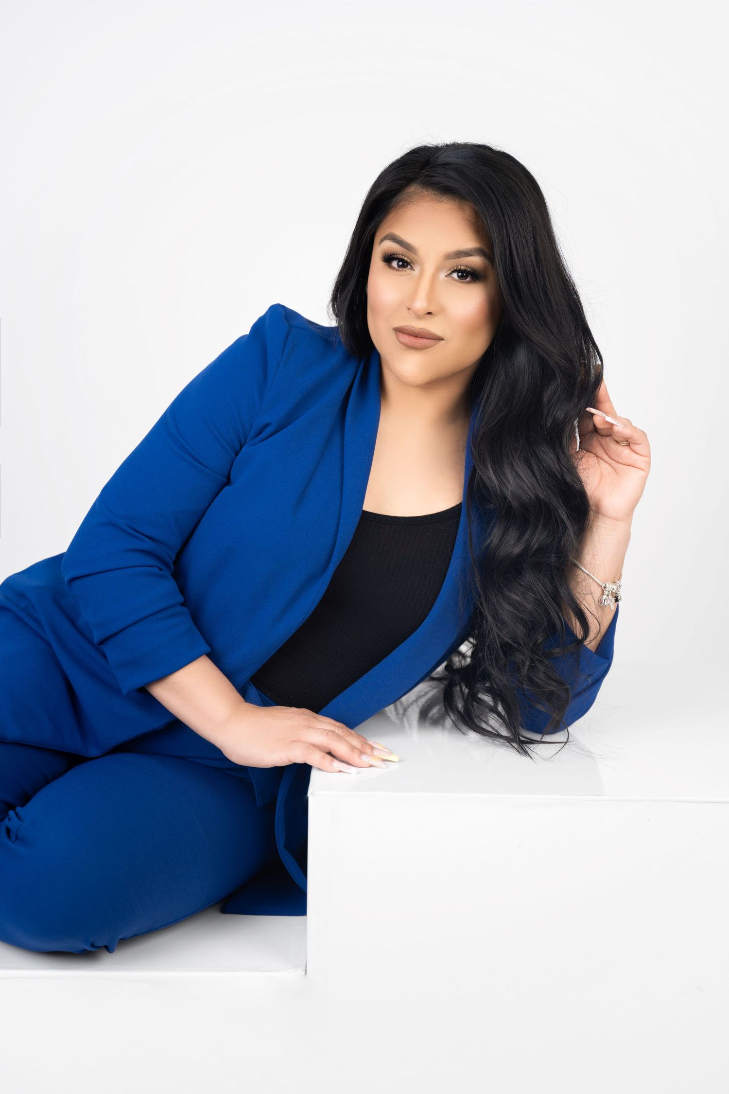 Woman with long wavy black hair poses on a white platform in a royal blue blazer and matching pants over a black top.