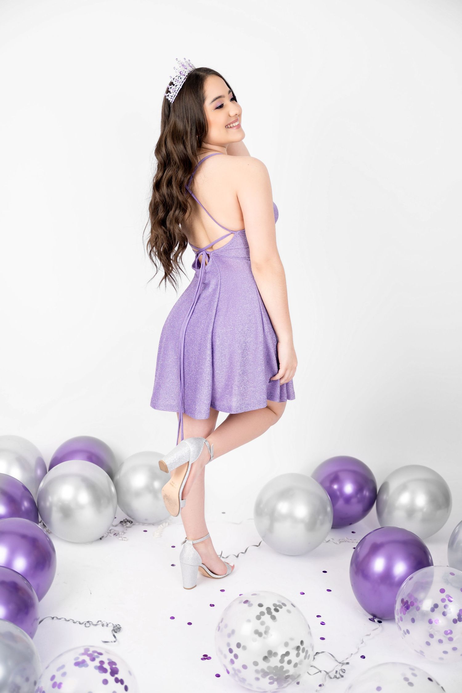 Smiling young woman in a purple sparkly dress and tiara poses among purple, silver and confetti balloons in a bright studio.