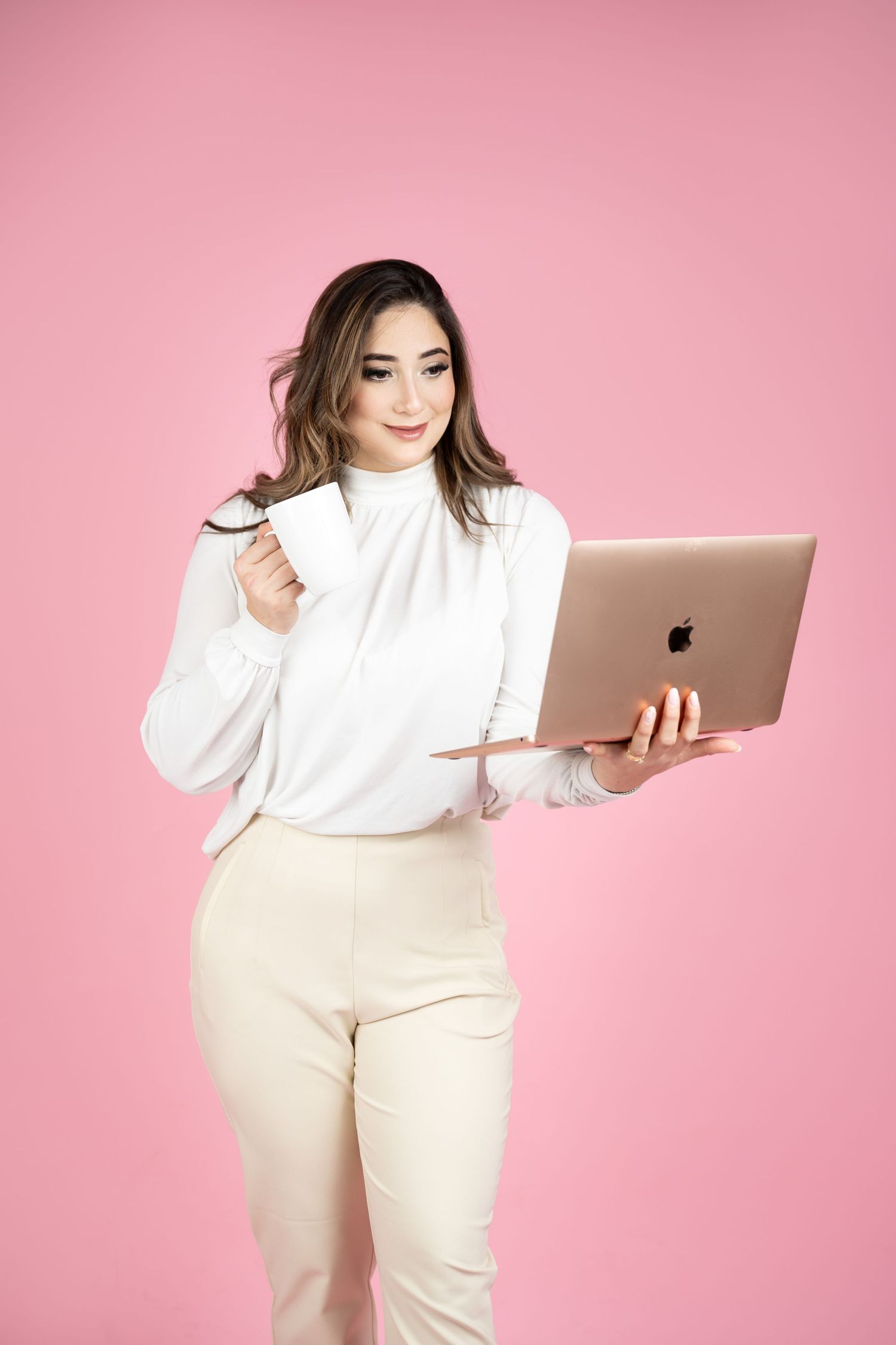 Young woman in white blouse holding rose gold MacBook laptop and coffee cup against pink background.