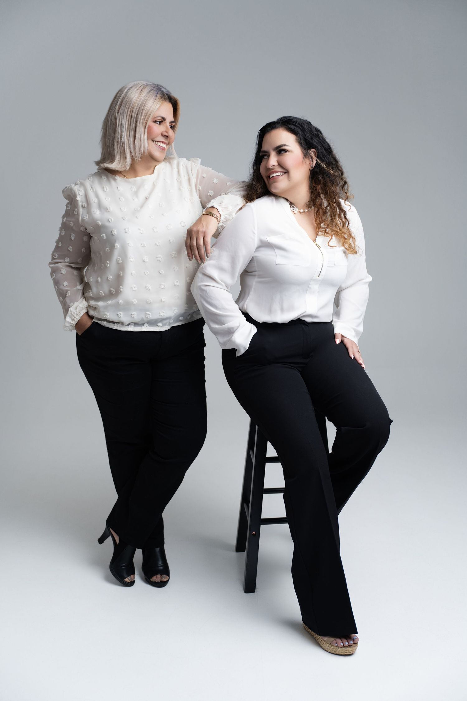 Two plus-size women in white blouses and black pants pose confidently in a studio setting.