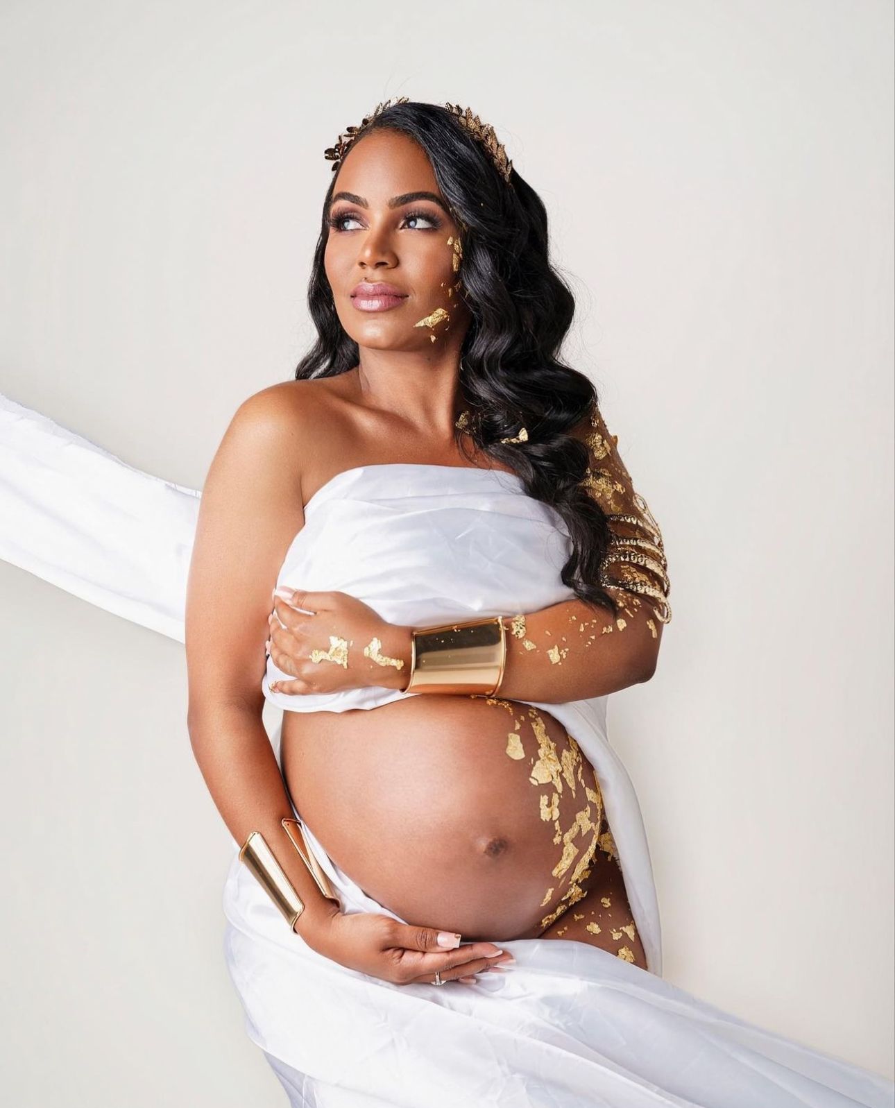 Pregnant woman in elegant white and gold maternity gown with crown, cradling baby bump in a professional photo shoot.