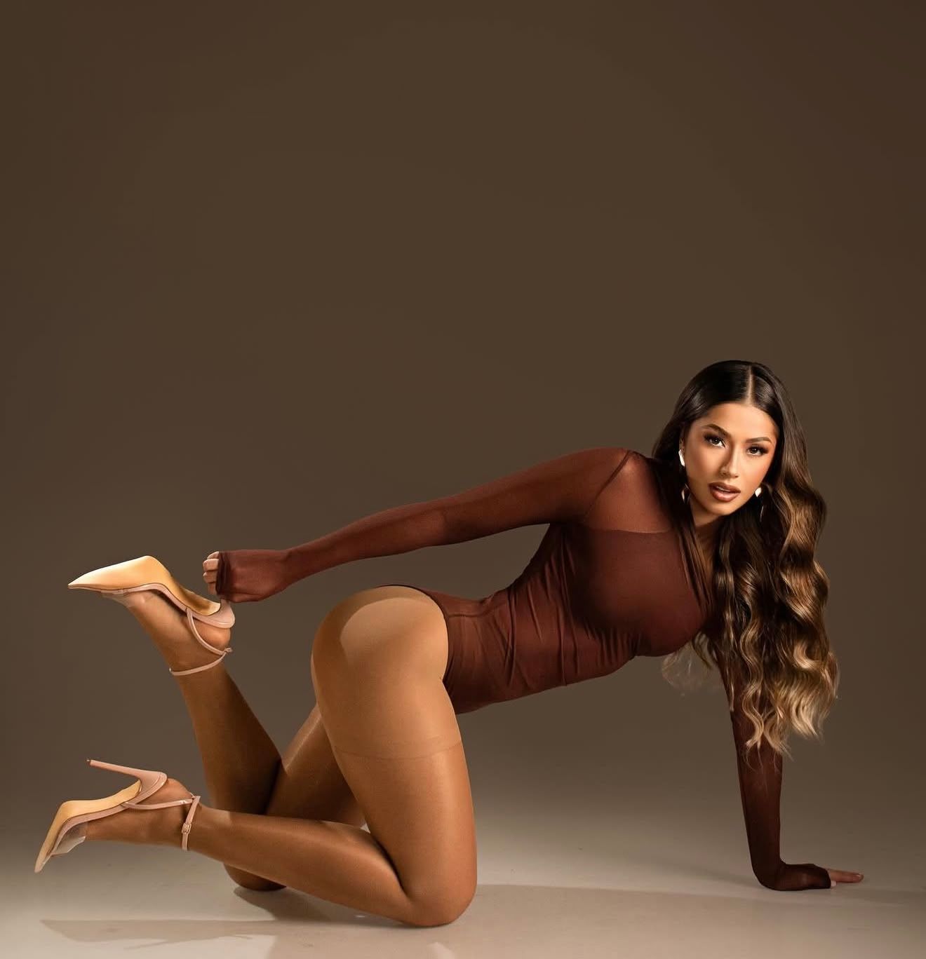 Woman in brown bodysuit and nude heels posing on floor with long wavy hair against brown background.