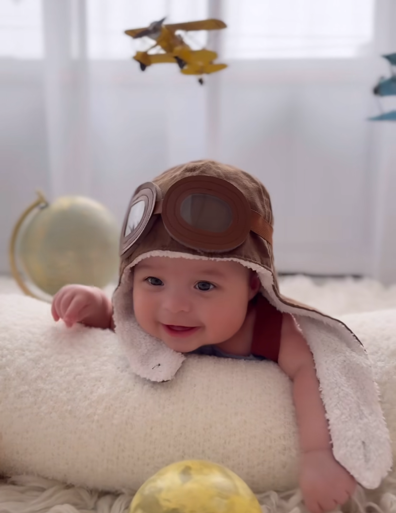 Smiling baby wearing a brown aviator hat with goggles, lying on a fluffy pillow with a toy airplane in the background.