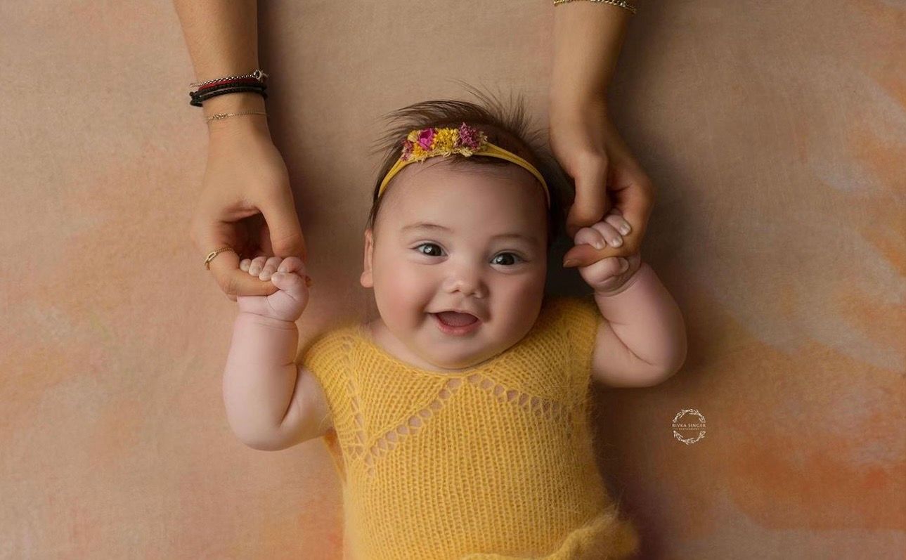 Smiling baby in yellow knit romper with floral headband, held by adult hands, lying on warm terracotta background.