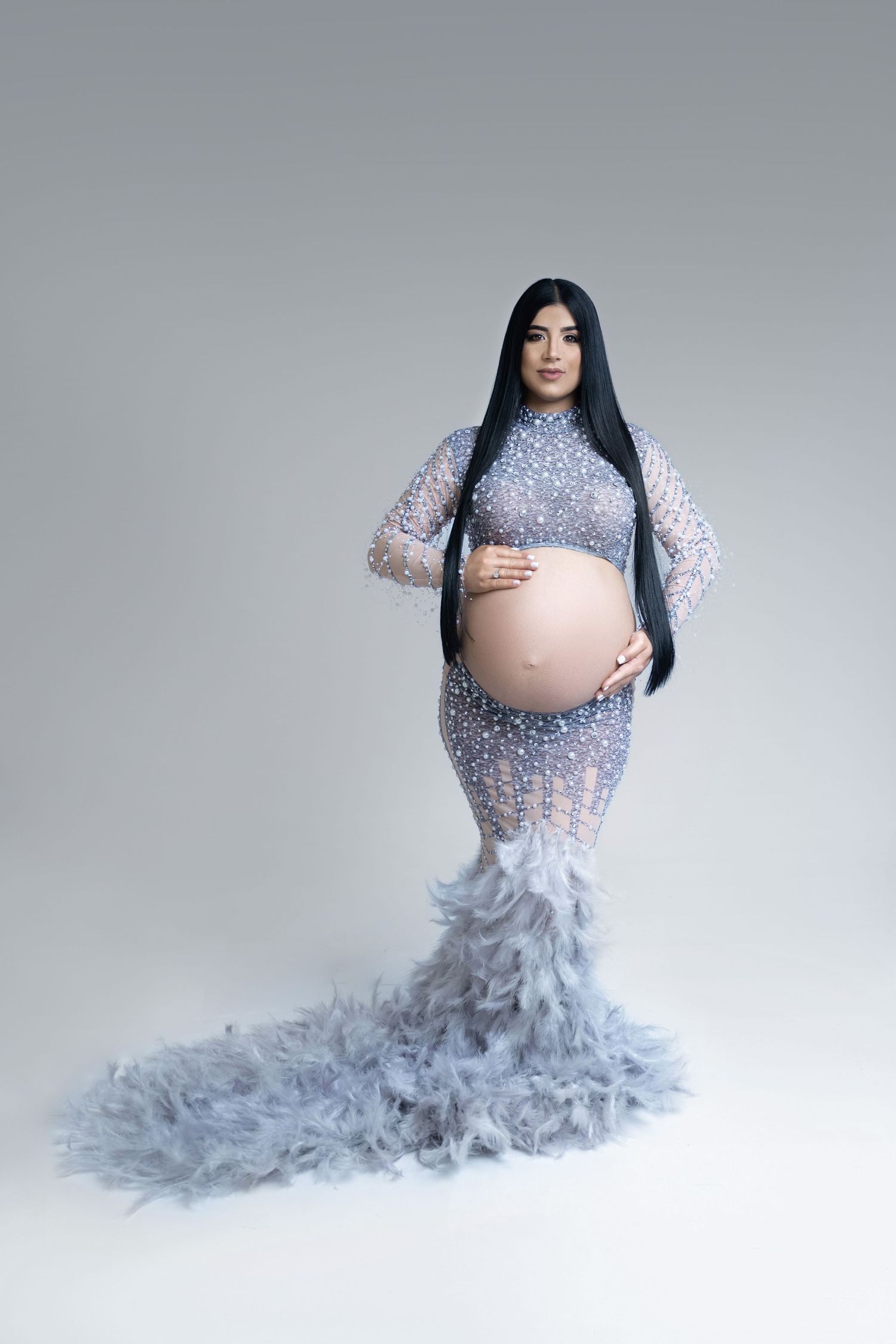 A maternity photoshoot featuring a grey feathered gown against a white backdrop.