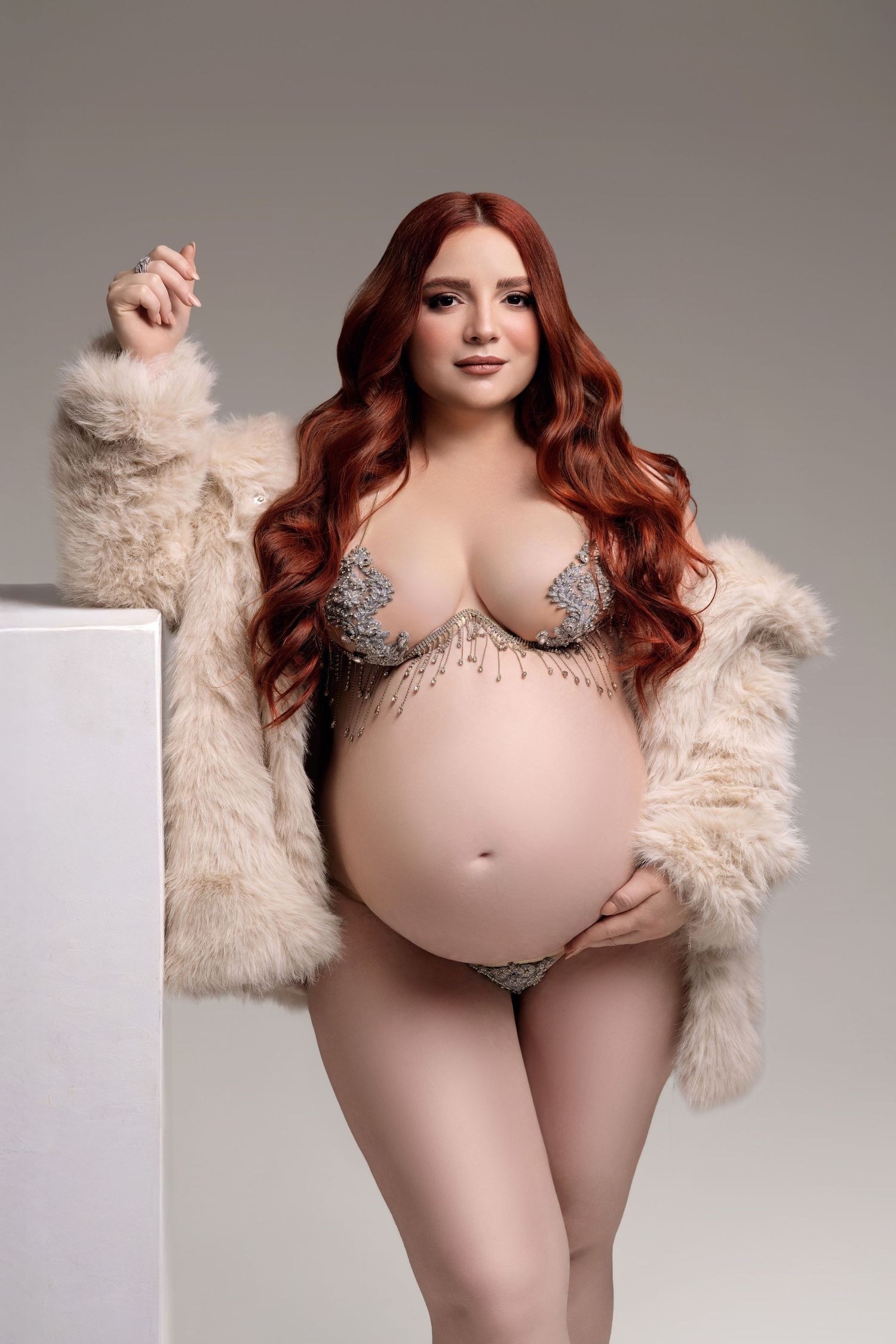 A model in a beige bodysuit and fur jacket poses against a gray backdrop in a fashion photoshoot.
