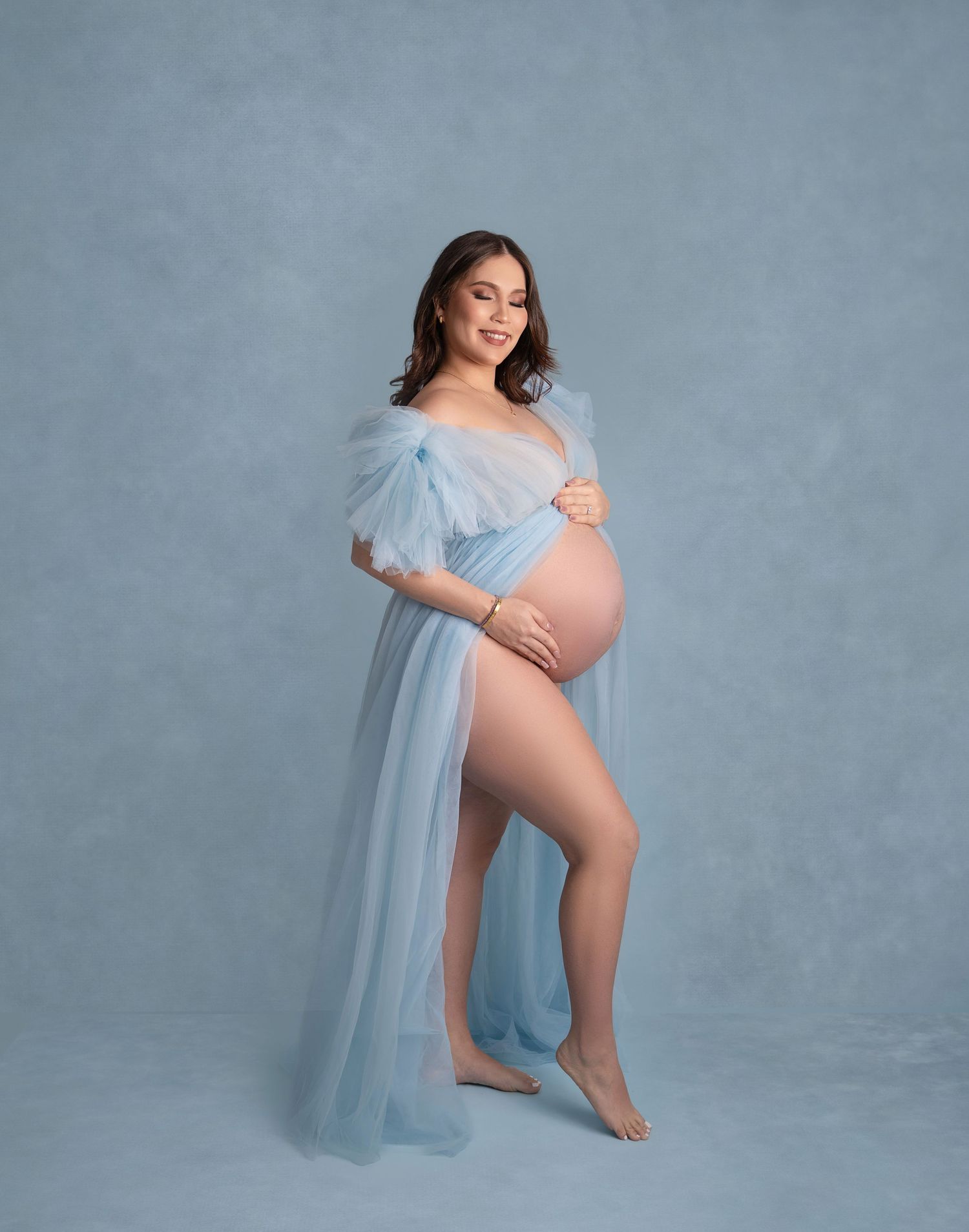 Maternity photo series showing a woman in a flowing blue tulle dress against a gray backdrop in artistic poses.