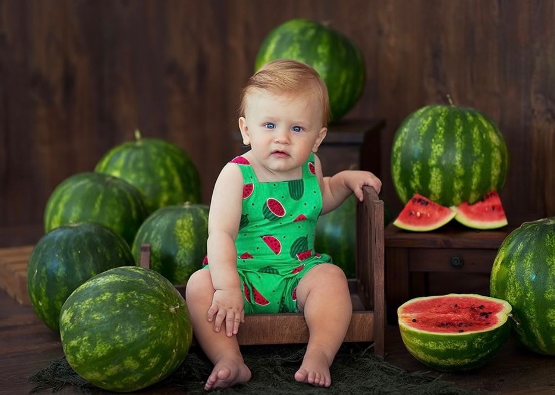 A child in a green outfit sits among large watermelons with sliced watermelon pieces displayed on a wooden floor.