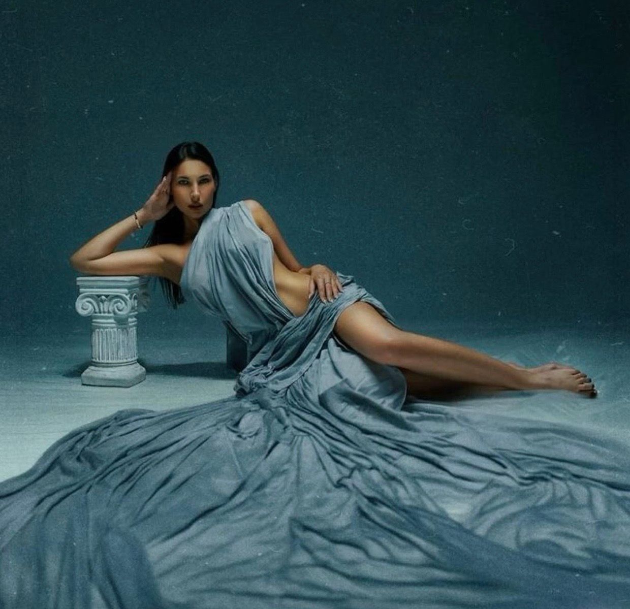 A woman in a flowing blue-gray gown reclines gracefully on the floor beside a white Greek column.