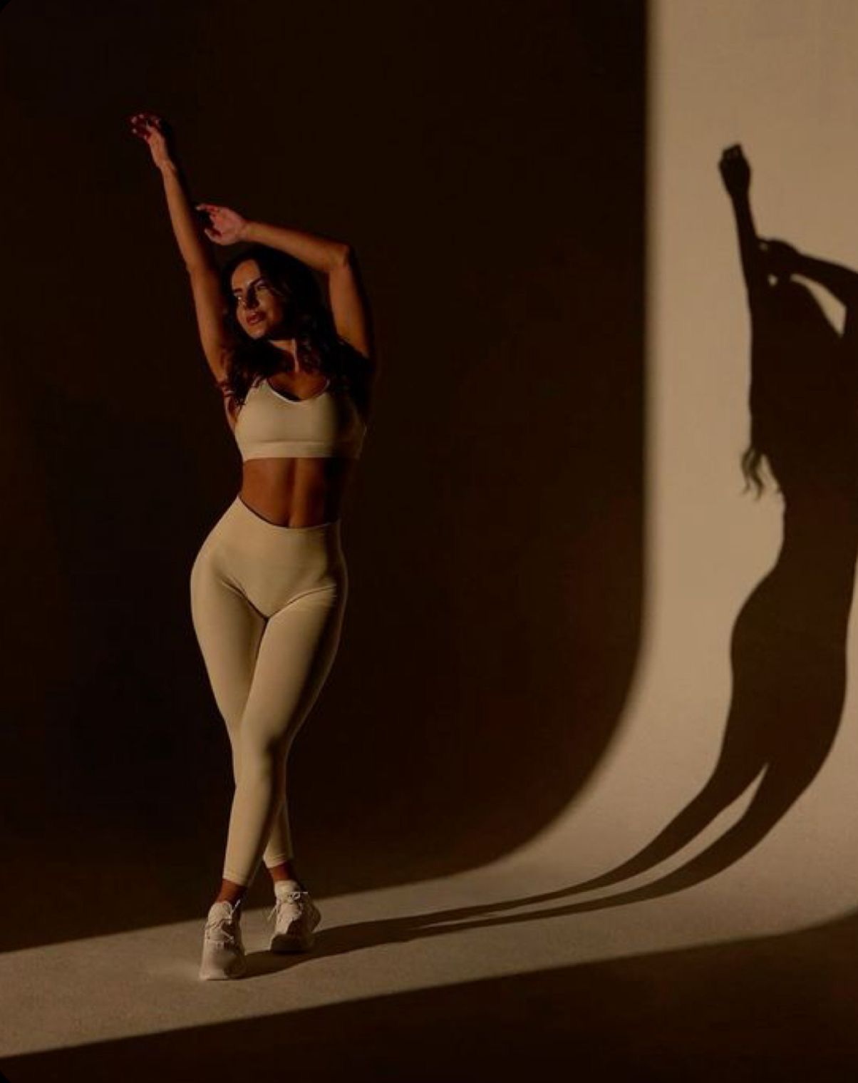 Woman in cream sports bra and leggings poses gracefully against a warm-lit wall with dramatic shadow.