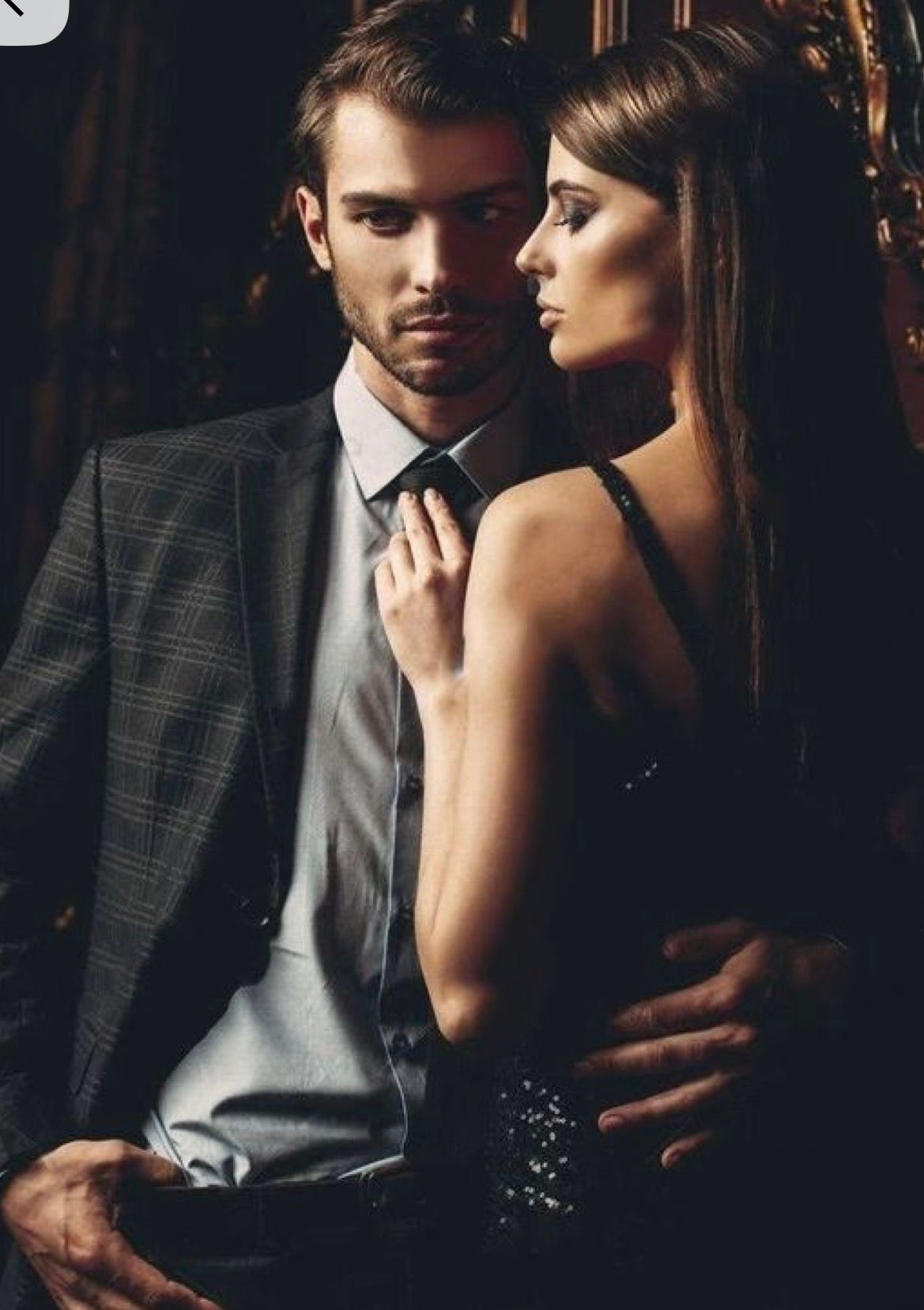 Elegant couple in formal attire, man in suit and tie with woman in black dress posing intimately in dark luxury setting.