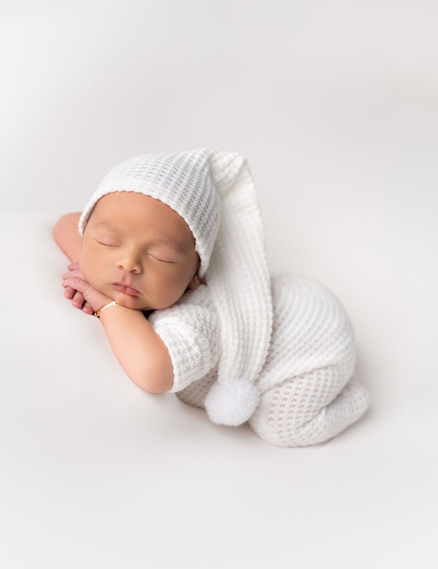 Sleeping newborn wearing white knitted hat and romper on soft white backdrop in studio setting.