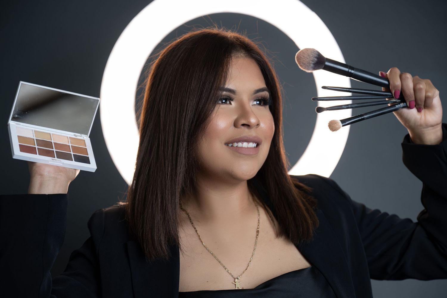 Makeup artist displaying cosmetic palette and brushes with ring light backdrop.
